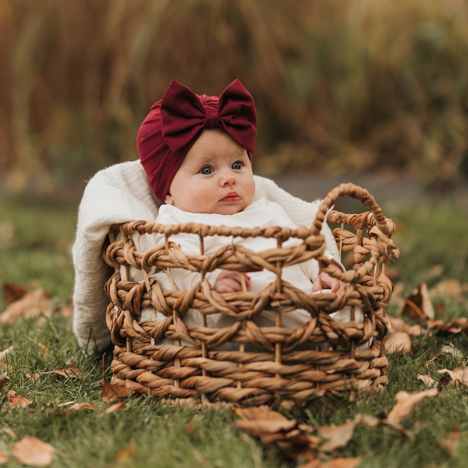 Infant sitting in a wicker basket outside in grass and leaves wearing the Bow Headwrap in Burgundy and white romper