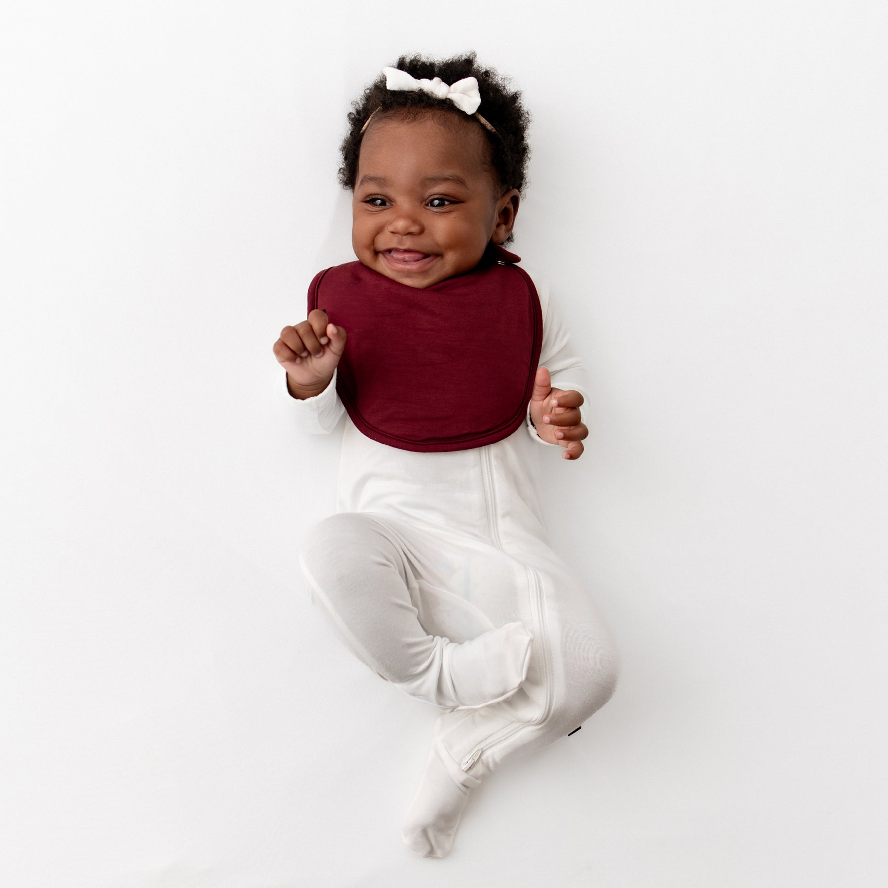 Smiling infant wearing the Cushy Bib in Burgundy overtop a Cloud zippered footie