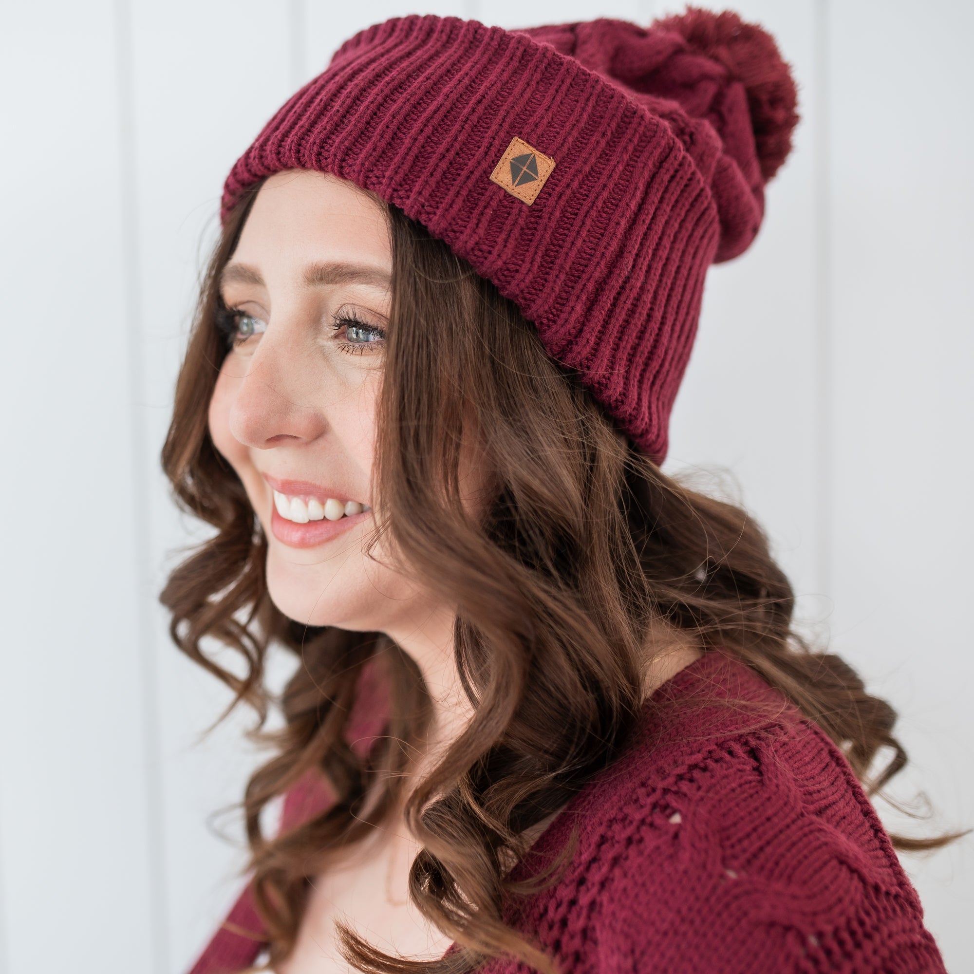 Cable Knit Adult Pom Beanie in Burgundy with the Kyte Baby vegan leather tag shown on a female model