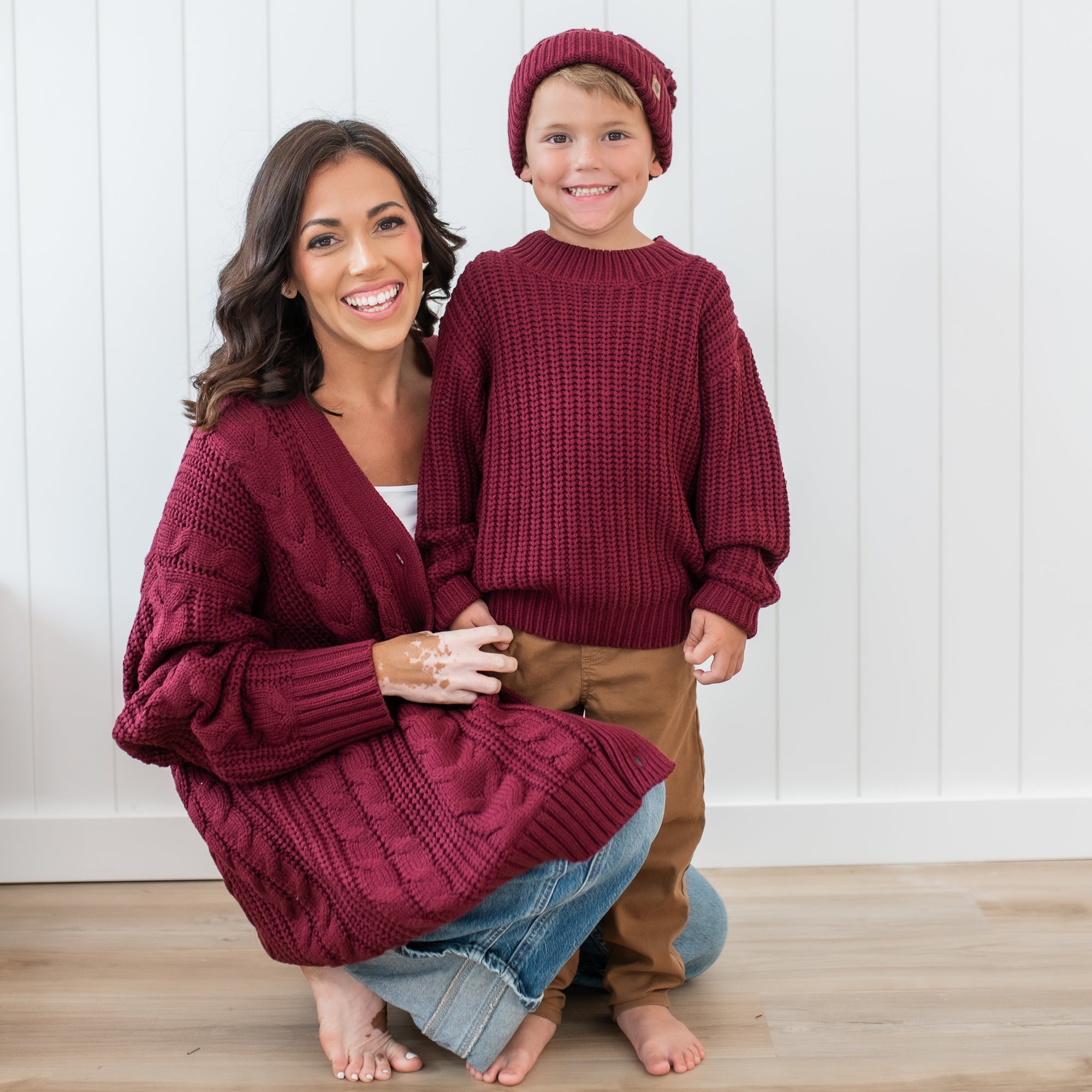 Mother and son wearing Burgundy Chunky knit items in front of a white paneled wall. Pictured is the Chunky Knit Sweater in Burgundy, Chunky cable knit beanie and Women's Cable Knit Cardigan