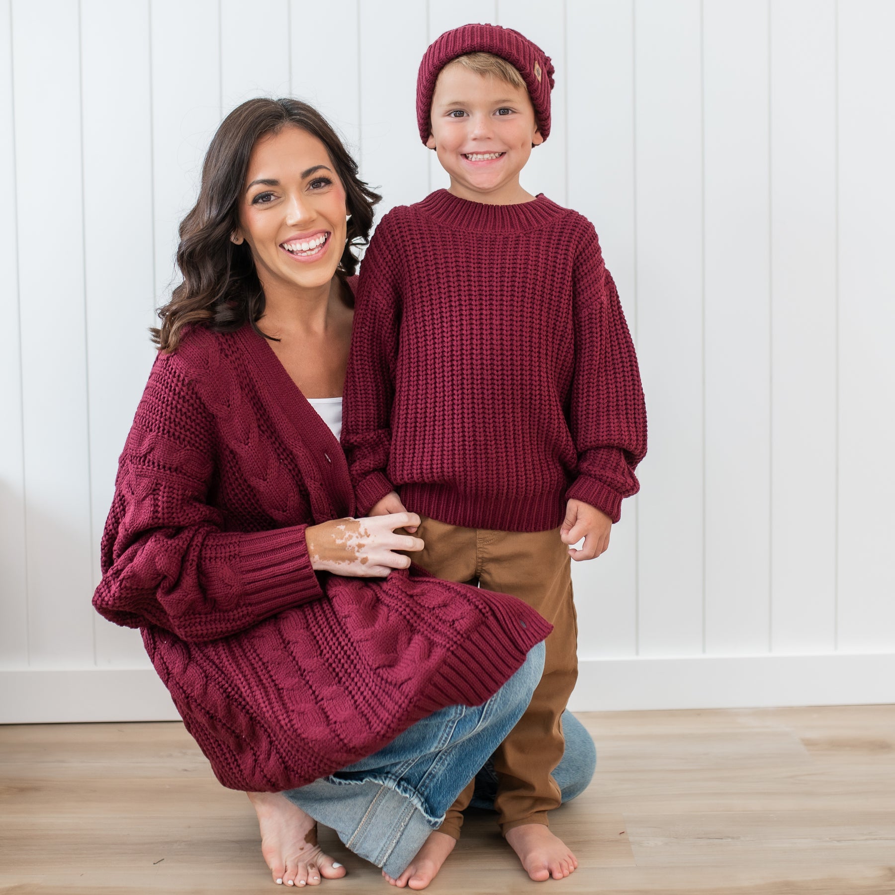 Mother and son wearing Burgundy Chunky knit items in front of a white paneled wall. Pictured is the Chunky Knit Sweater in Burgundy, Chunky cable knit beanie and Women's Cable Knit Cardigan