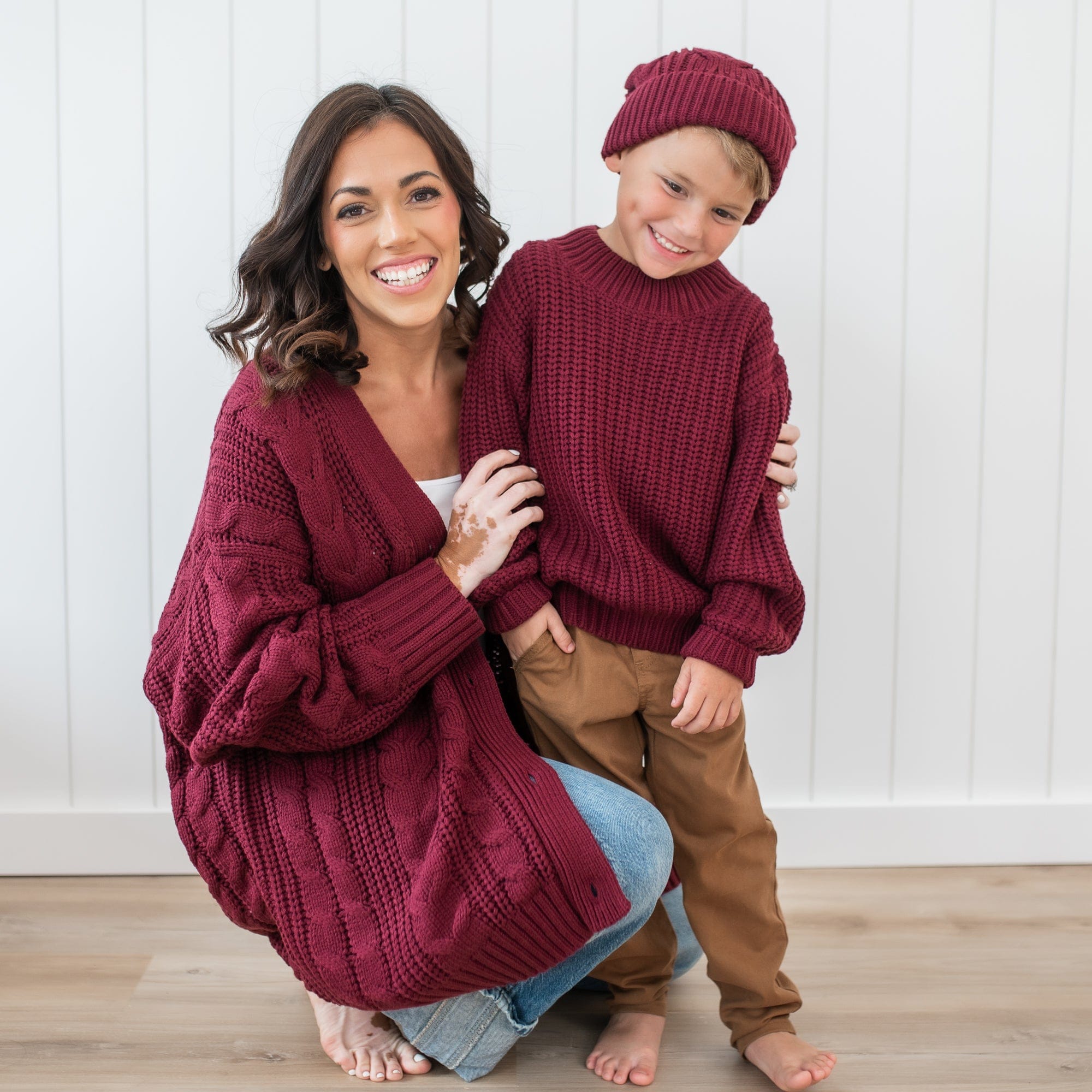 Mother, wearing the Cable Knit Adult Cardigan in Burgundy, kneeling beside her son who is matching in a Chunky knit sweater