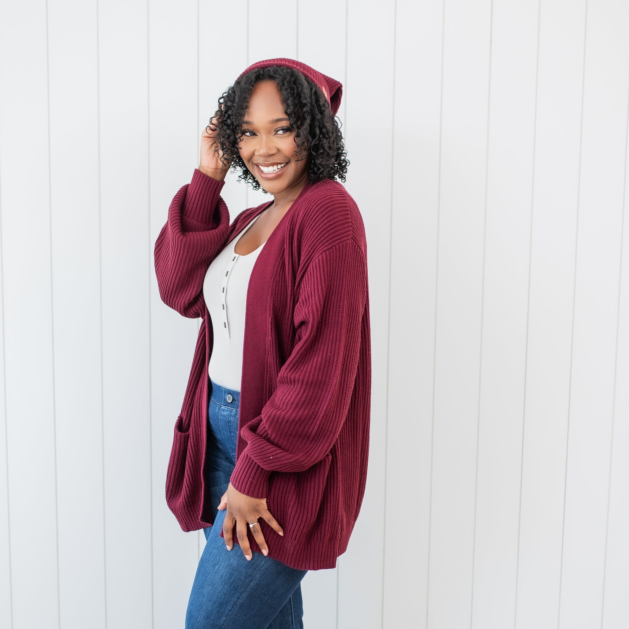 Female standing in front of a white paneled wall wearing the Chunky Knit Adult Pom Beanie in Burgundy and matching chunky knit women's oversized cardigan