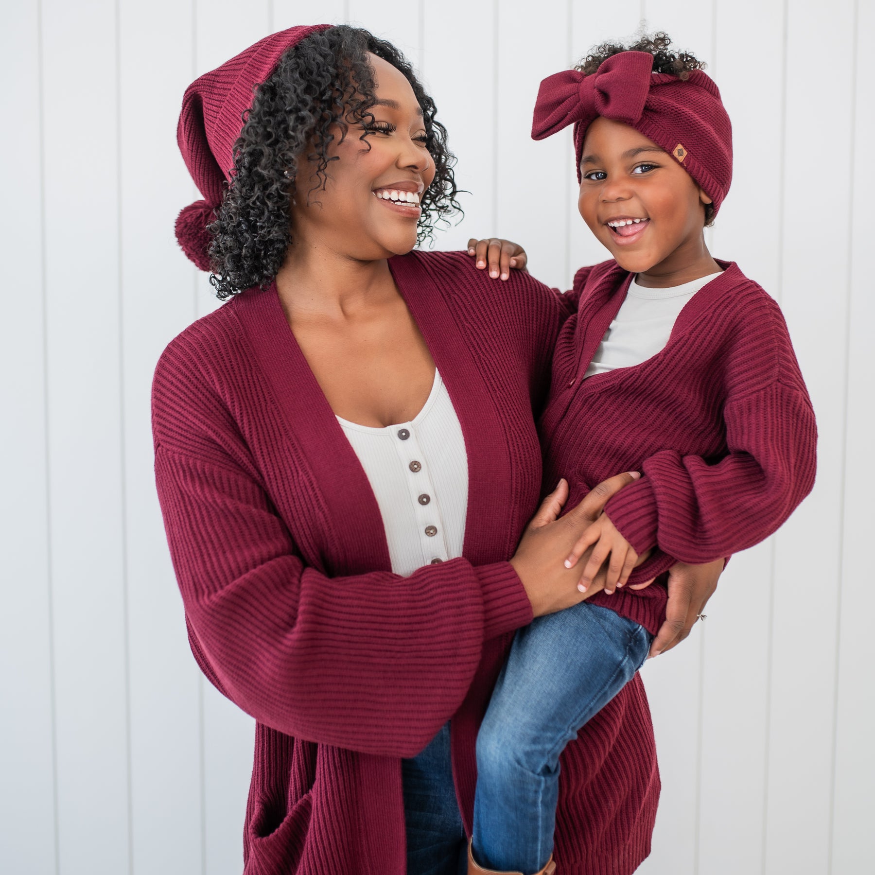 Mother holding daughter standing in front of  a white paneled wall wearing various Burgundy Chunky Knit items