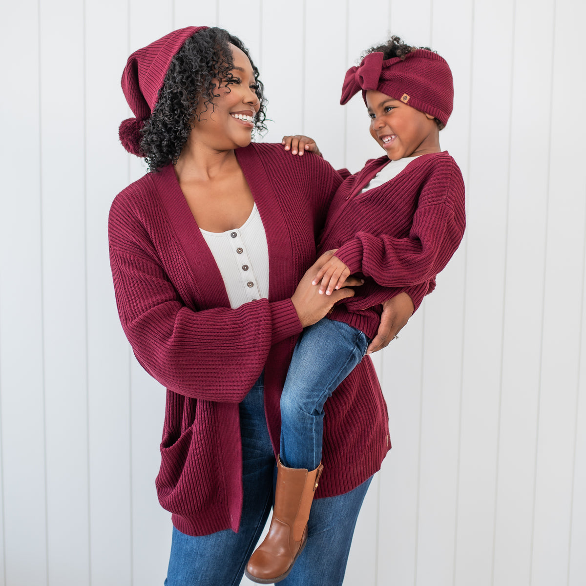Mother wearing the Chunky Knit Women's Oversized Cardigan in Burgundy holding her daughter who is matching in Burgundy Chunky cardigan and chunky knit bow headband
