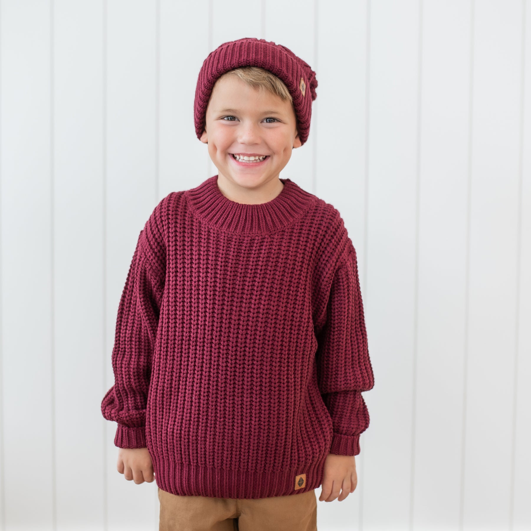Smiling boy wearing the Chunky Knit Sweater in Burgundy and Chunky Cable knit beanie standing in front of a white paneled wall