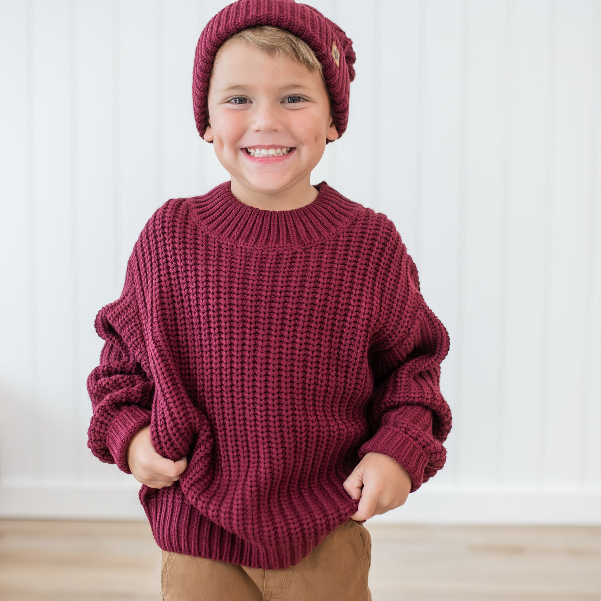 Smiling young boy wearing the Chunky Knit Sweater in Burgundy with dark tan colored pants