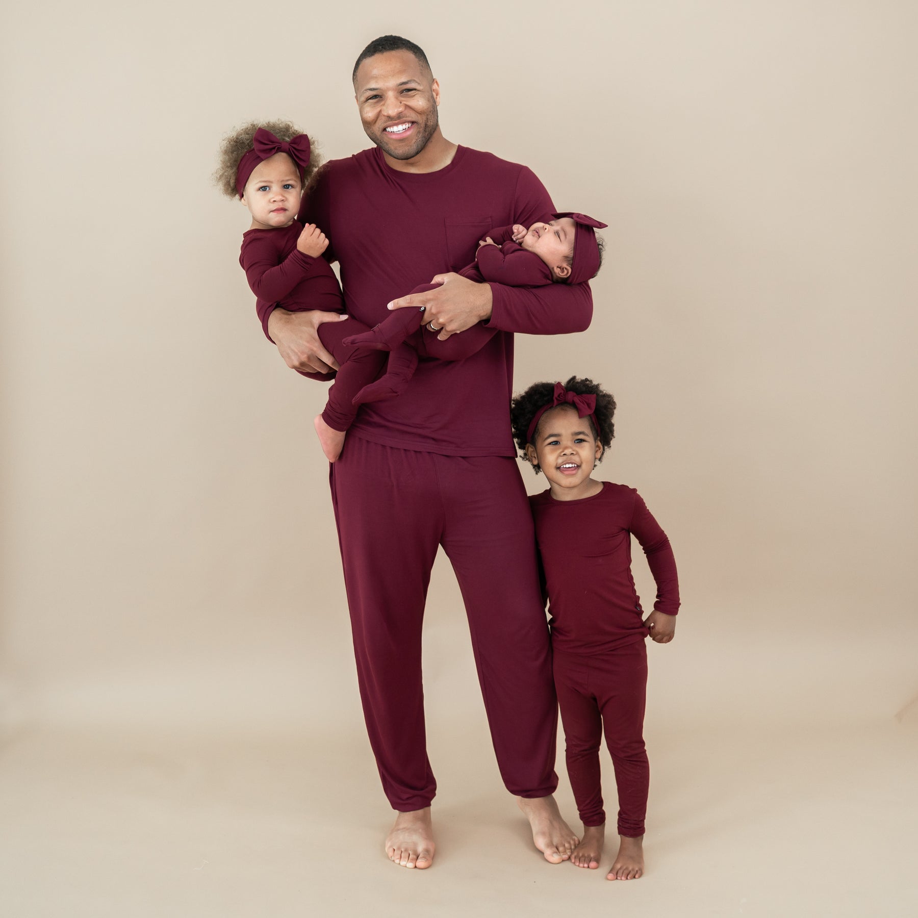 Family matching in Burgundy. Father holding younger two daughters with his oldest daughter standing right next to him. Pictured are the men's joggers, zippered footie and toddler pajama sets