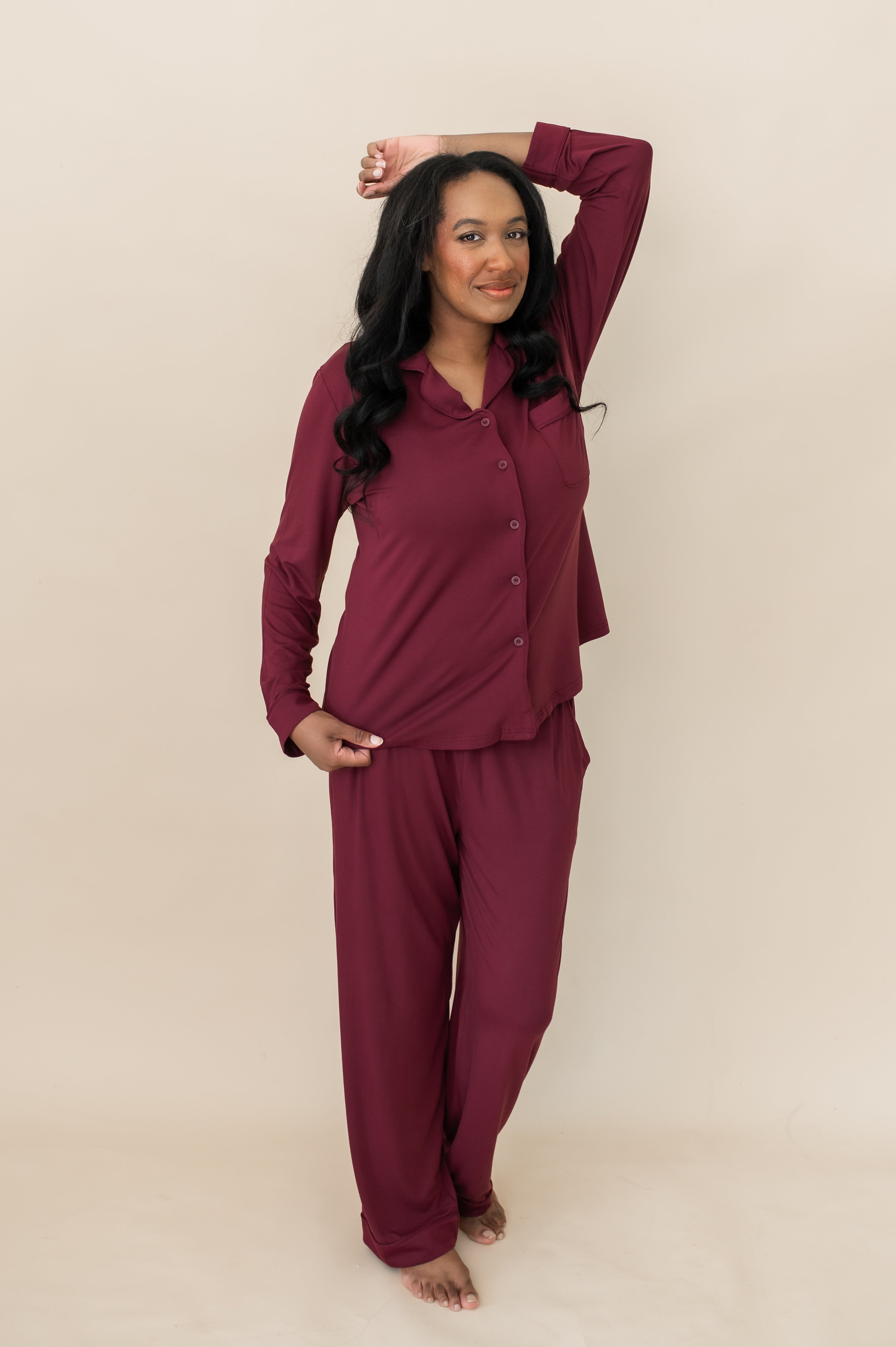 Female modeling the Long-Sleeved Women's Pajama Set in Burgundy with Burgundy Trim with on arm resting on the top of her head and the other hand holding the bottom hem on the shirt