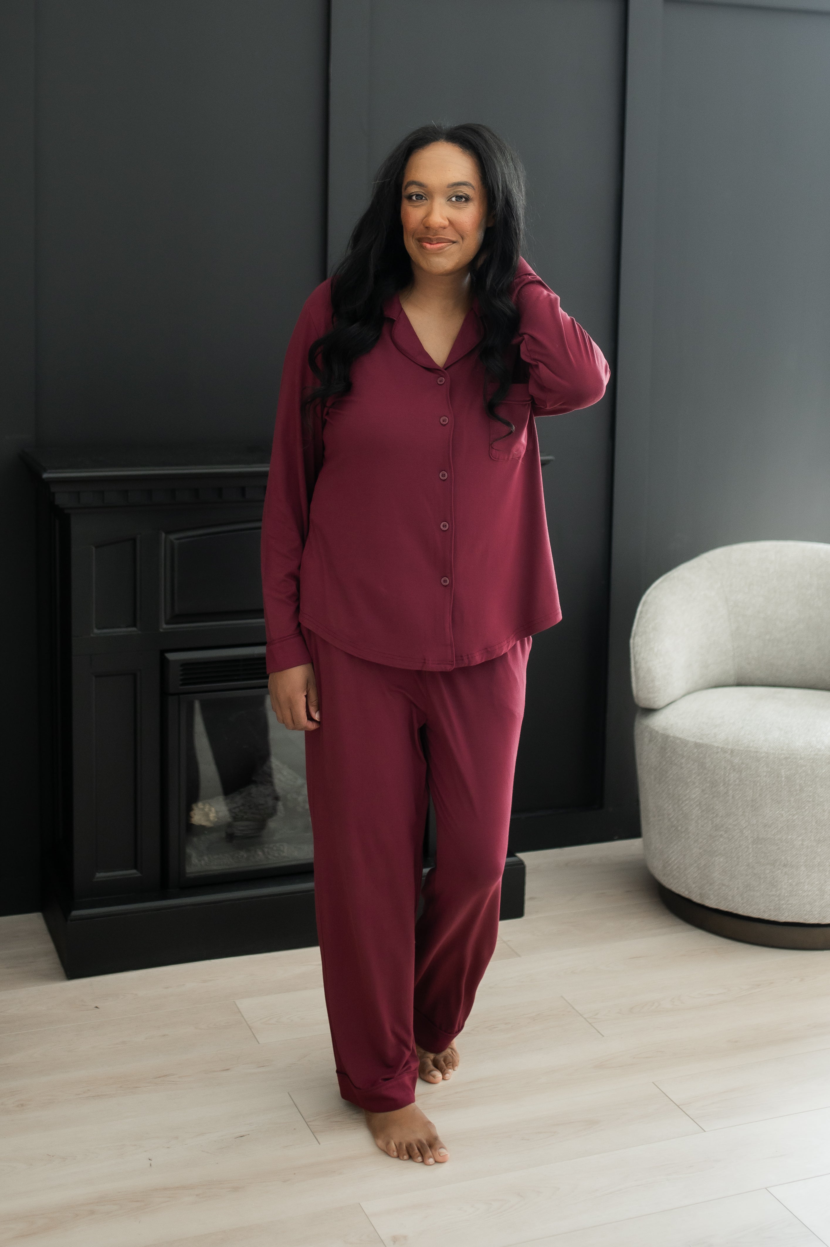 Female wearing the Long-Sleeved Women's Pajama Set in Burgundy with Burgundy Trim standing in front of a black wall and black fireplace