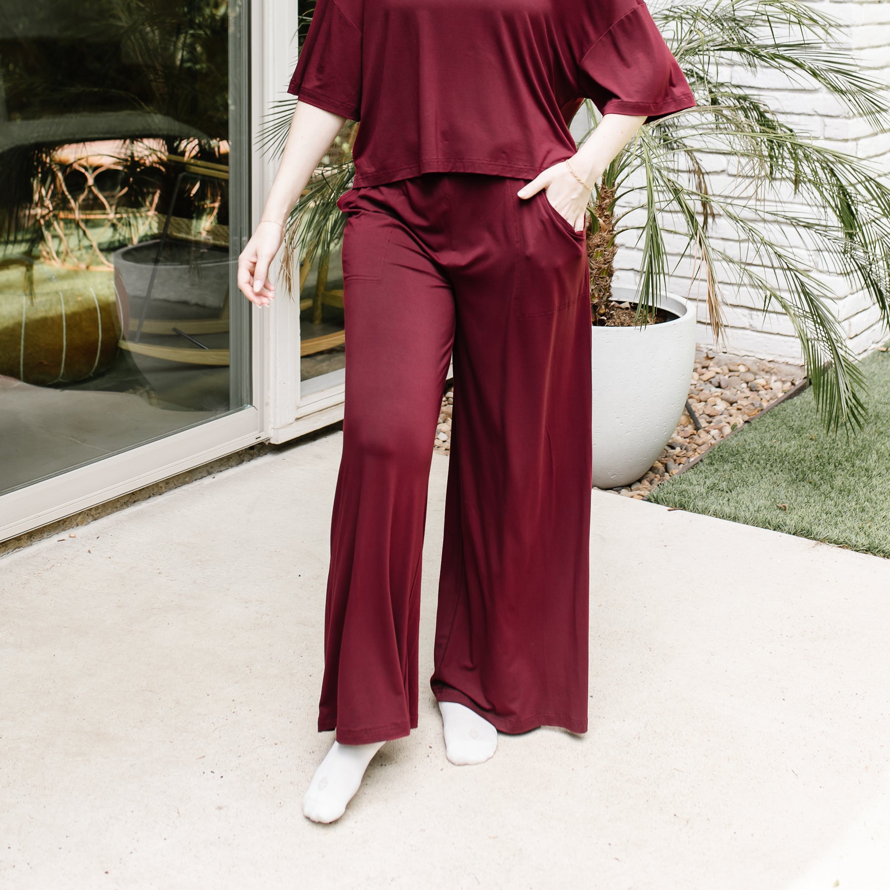 A model standing on a sidewalk in front of a white brick house wearing the Women's Wide Leg Pant in Burgundy