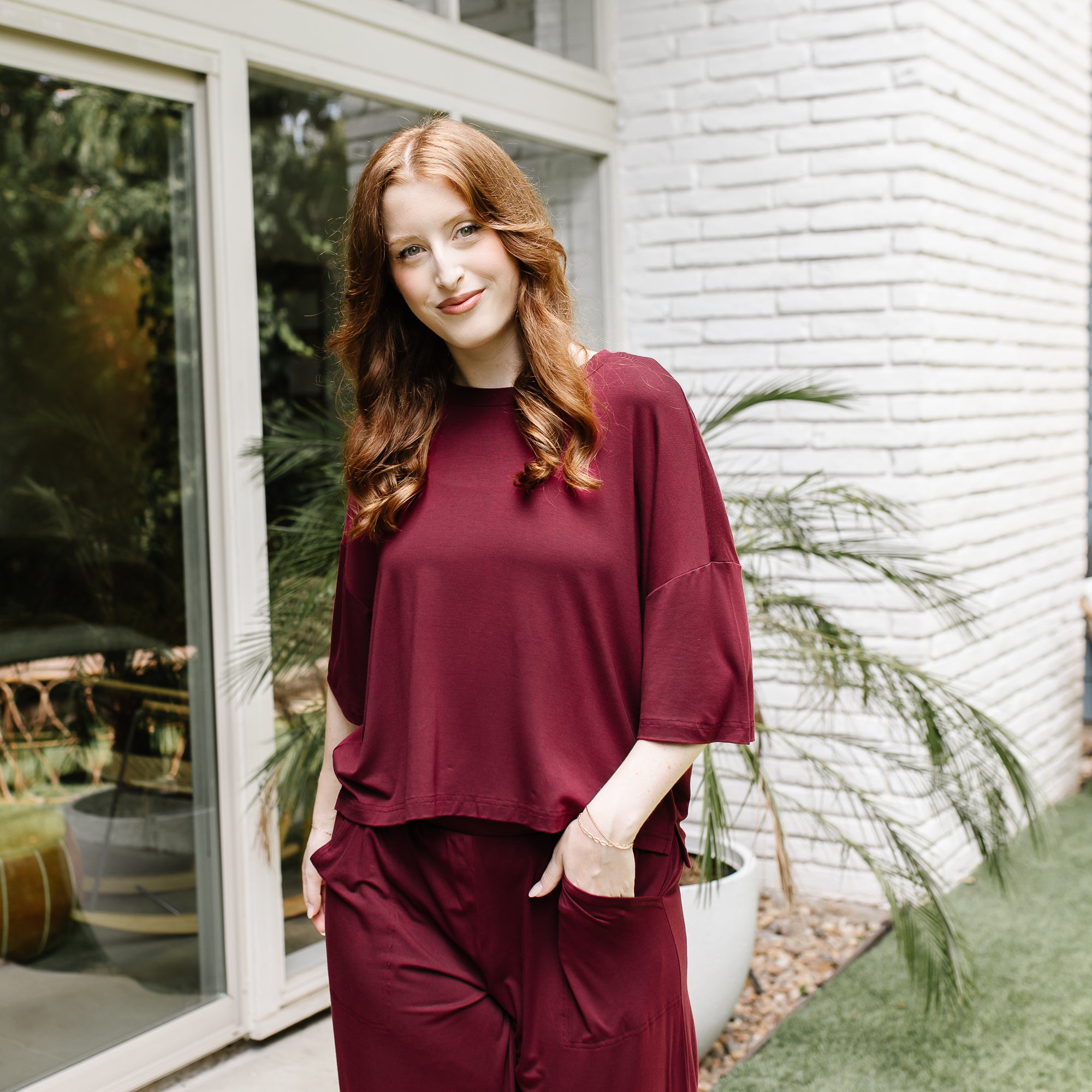 Female model standing in front of a white brick house wearing the Women's Short Sleeve Tee in Burgundy with one hand in the pocket of the matching wide let pant