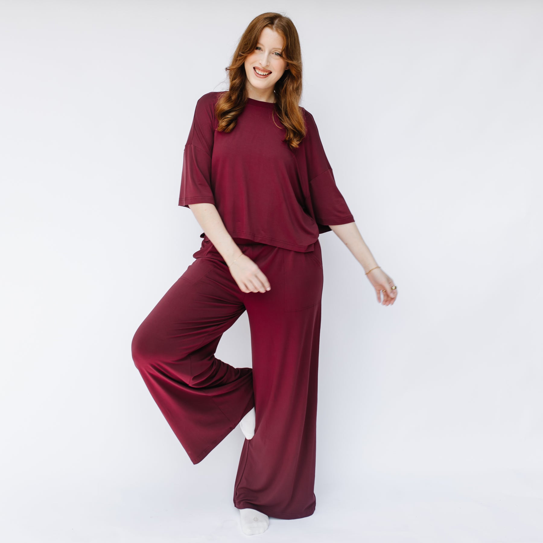 Smiling female standing in front of a white wall wearing the Women's Short Sleeve Tee in Burgundy with matching wide leg pants