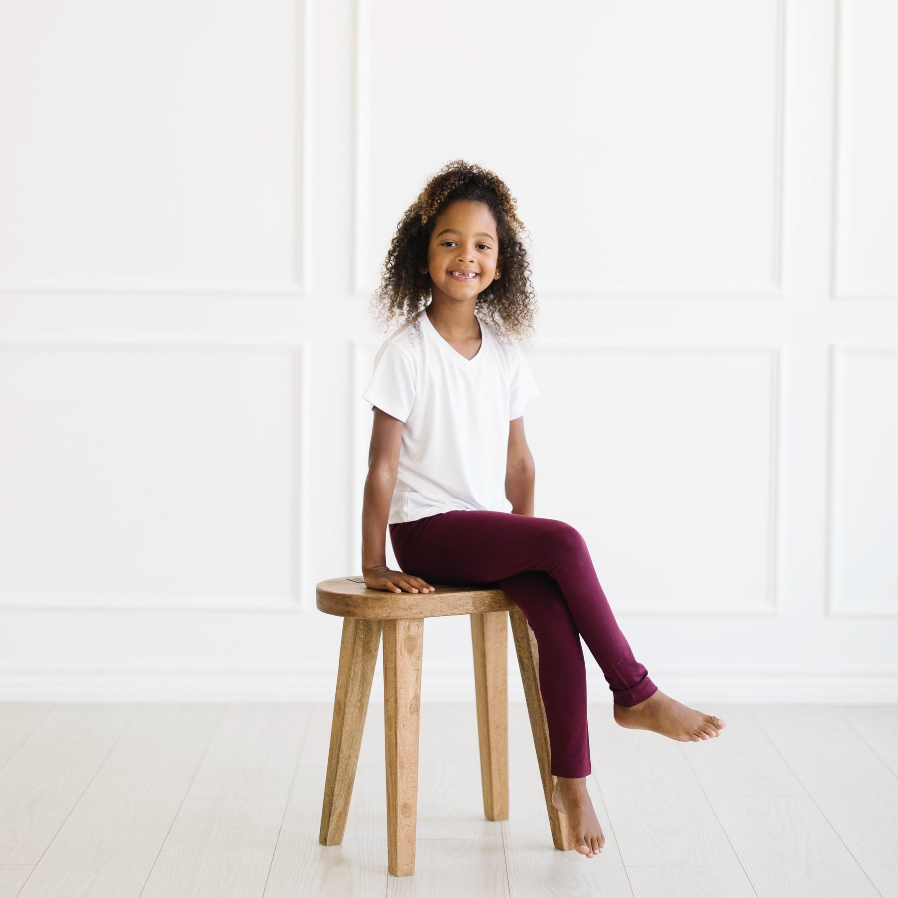 Young model sitting on a wooden stool wearing the Toddler Leggings in Burgundy paired with a Snow V-neck