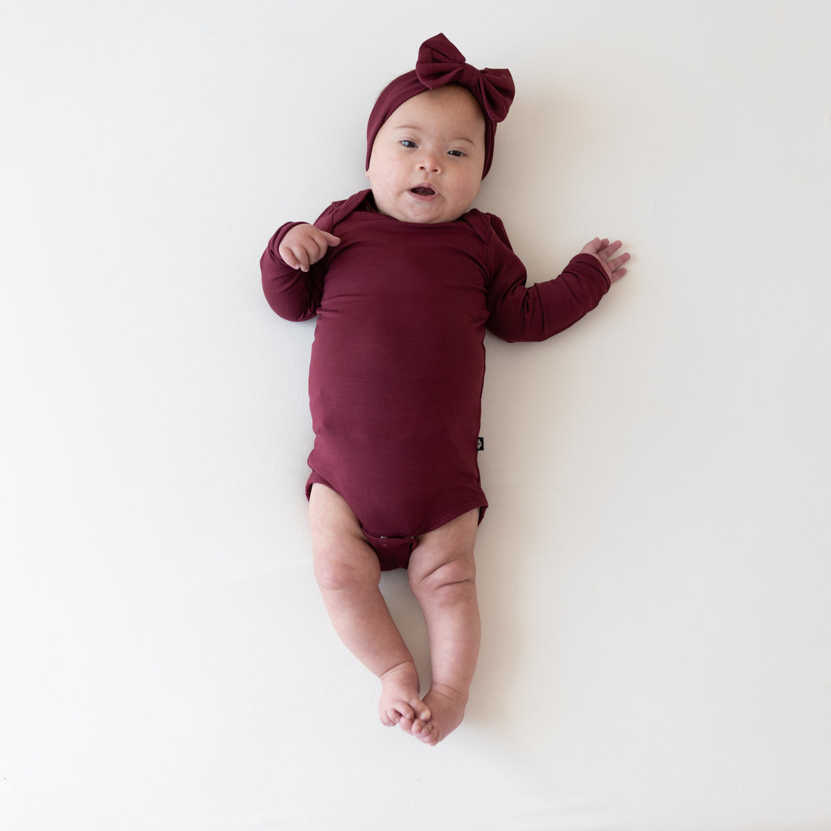 Infant girl laying wearing the Long Sleeve Bodysuit in Burgundy paired with matching long sleeve bodysuit