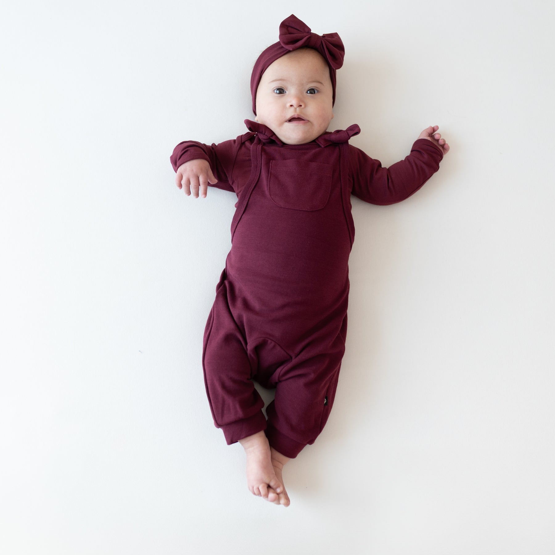 Infant staring straight into the camera wearing the Bamboo Jersey Overall in Burgundy paired with matching long sleeve bodysuit and bow headband on a light neutral background