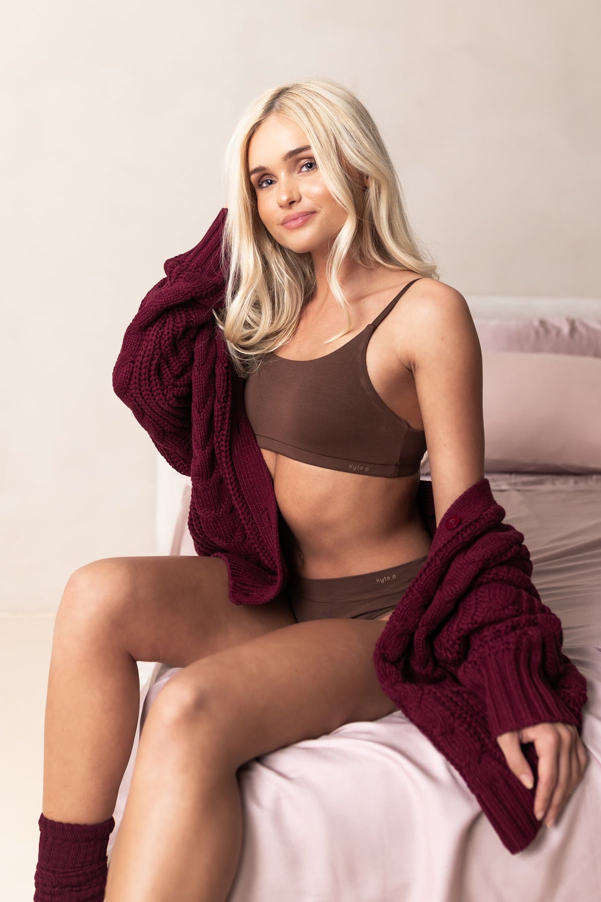 Woman in a brown scoop neck bralette and underwear set with a maroon cardigan on a light background