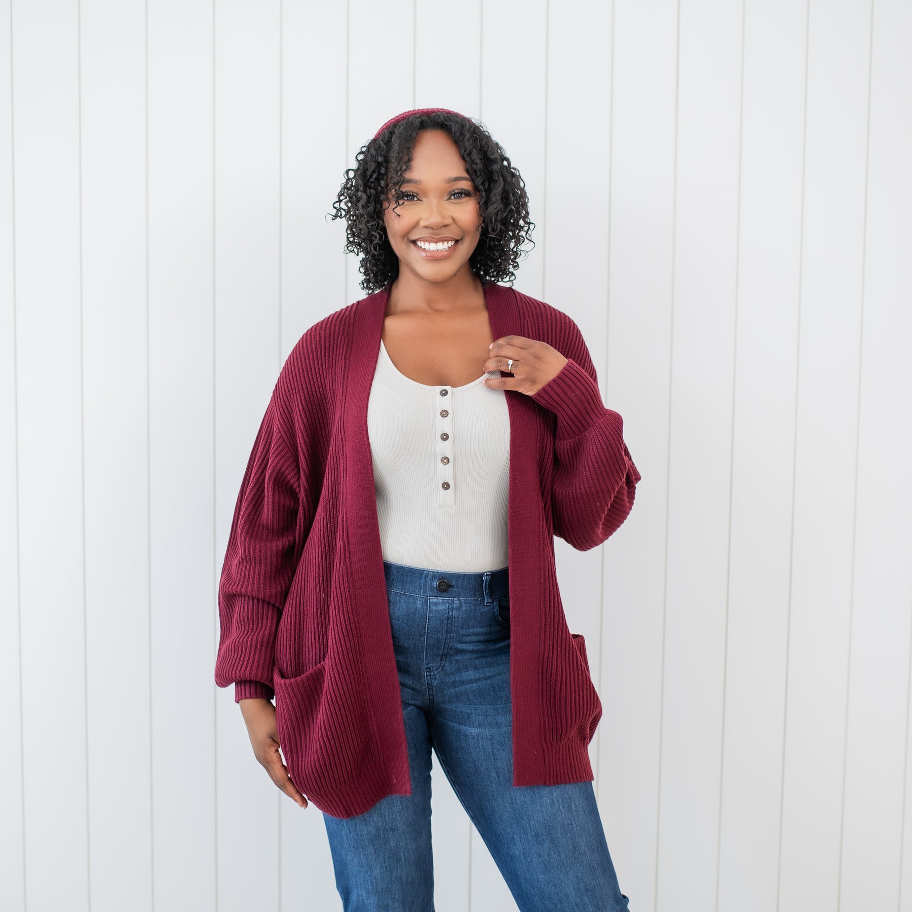 Female model wearing the Chunky Knit Women's Oversized Cardigan in Burgundy paired with medium wash jeans standing in front of a white paneled wall
