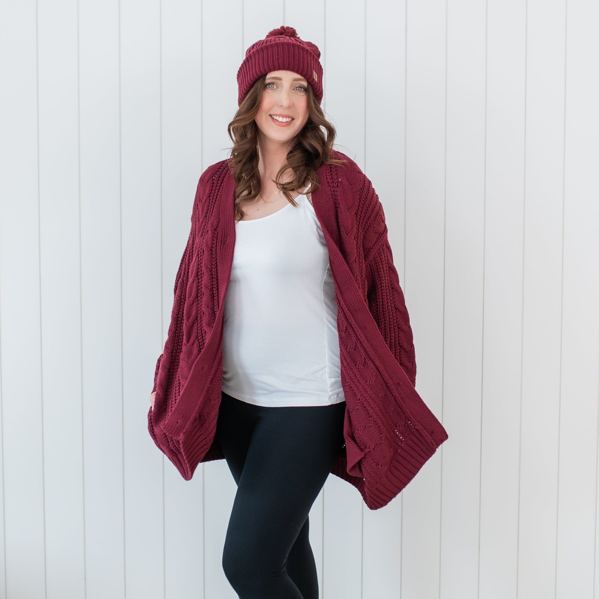 Female model wearing the Cable Knit Adult Cardigan in Burgundy and matching chunky knit beanie standing in front of a white paneled wall