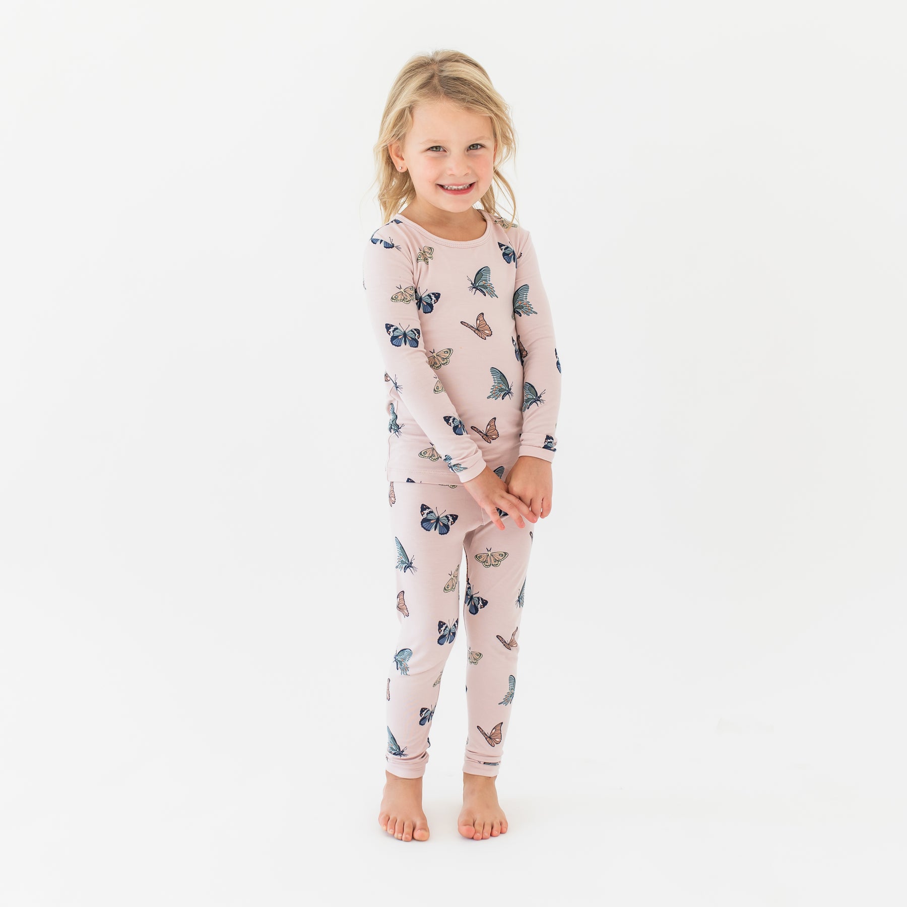 Young smiling girl standing with her hands clasped together wearing the Long Sleeve Pajamas in Blush Butterfly