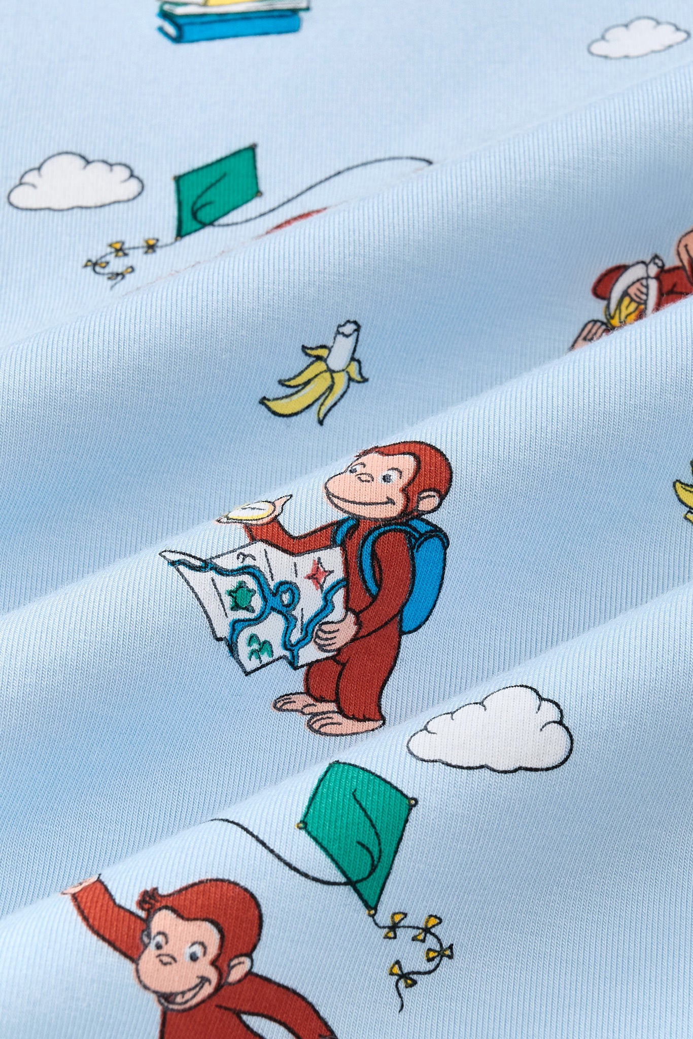 A repeating, all-over print featuring: Curious George sitting on a stack of books and reading a book with a red cover; Curious George eating a banana and leaning against a yellow hat; Curious George wearing a blue backpack and looking at a map and a compass; Curious George hanging onto the string of a green kite and soaring in the air; small white clouds; a cluster of three bananas; and a half-peeled, bitten banana, over a sky blue background color called Breeze.