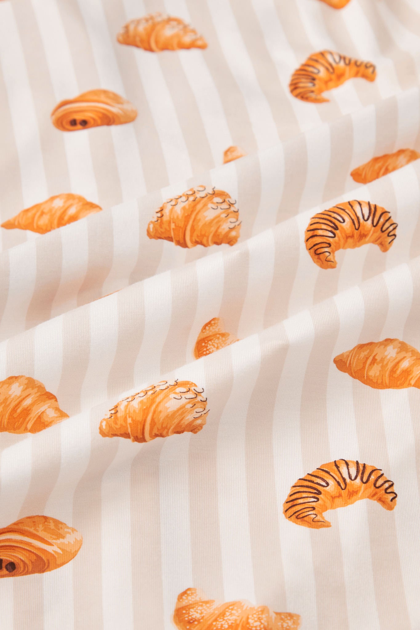 An all-over print of various types of croissants over a background of vertical stripes in white and a light beige color named Vanilla.