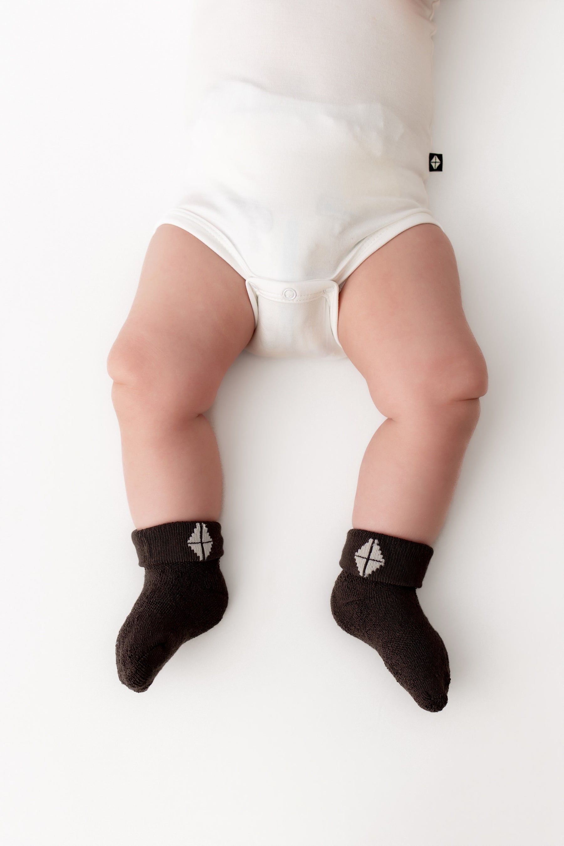 A baby lying on a white surface wearing white bodysuit bottoms and dark brown socks with a small white geometric logo near the cuff.