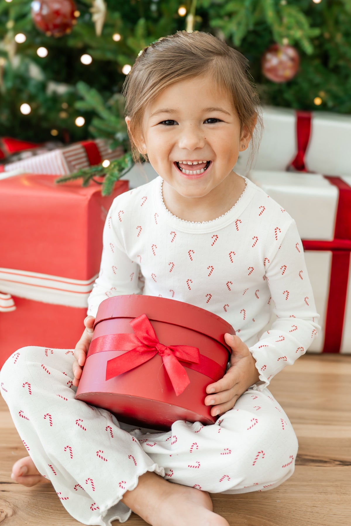 A young child wearing white holiday set with a red candy cane pattern sits on a wooden floor, smiling while holding a red gift box with a red ribbon bow. A Christmas tree and wrapped presents are blurred in the background.