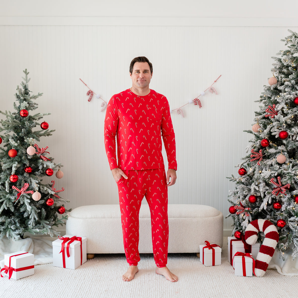 Male model standing in front of a cream ottoman between two decorated Christmas trees wearing the Men's Jogger Set in Candy Cane with one hand in the jogger pants pocket