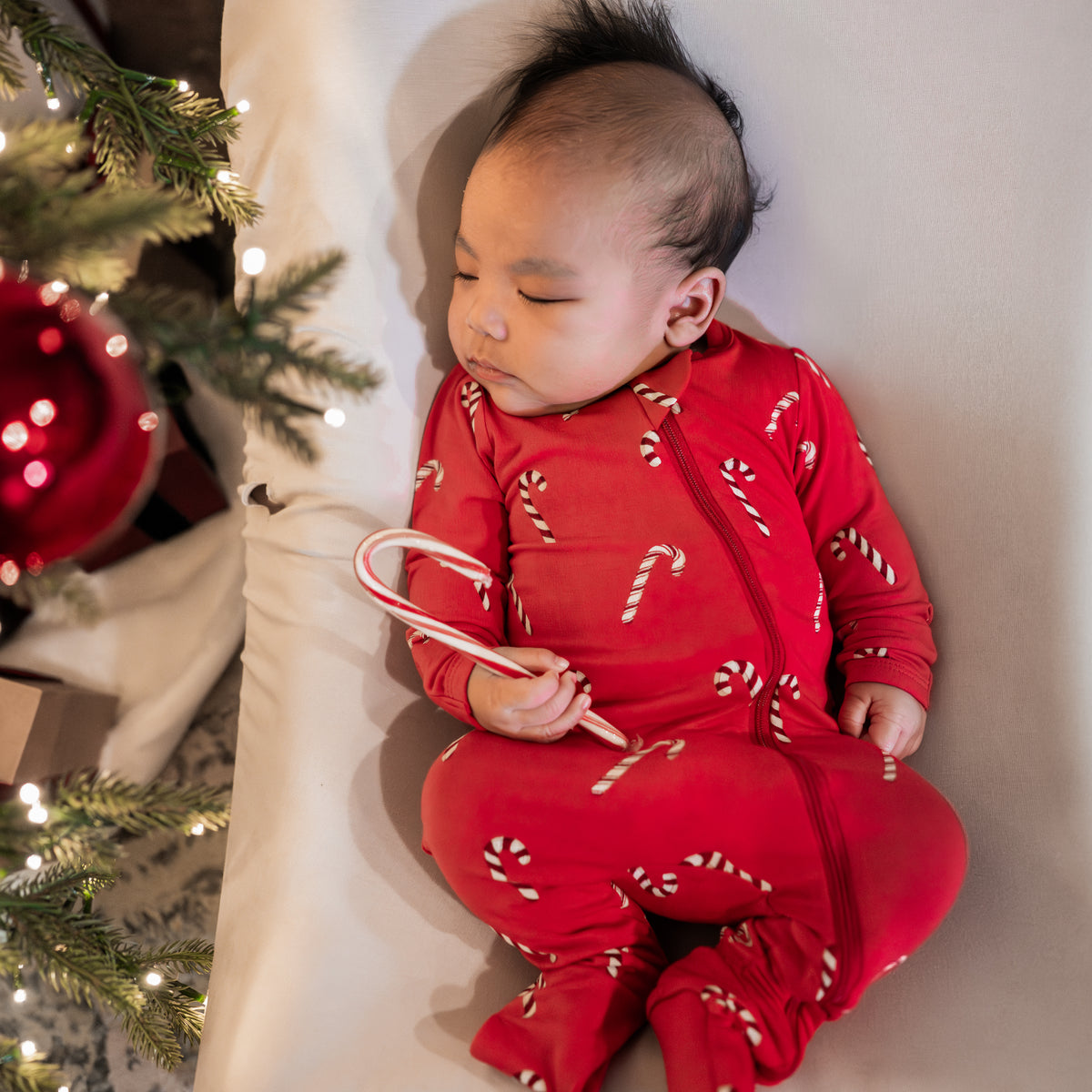Sleeping infant holding a Candy Cane wearing the Zippered Footie in Candy Cane laying beside a decorated Christmas Tree