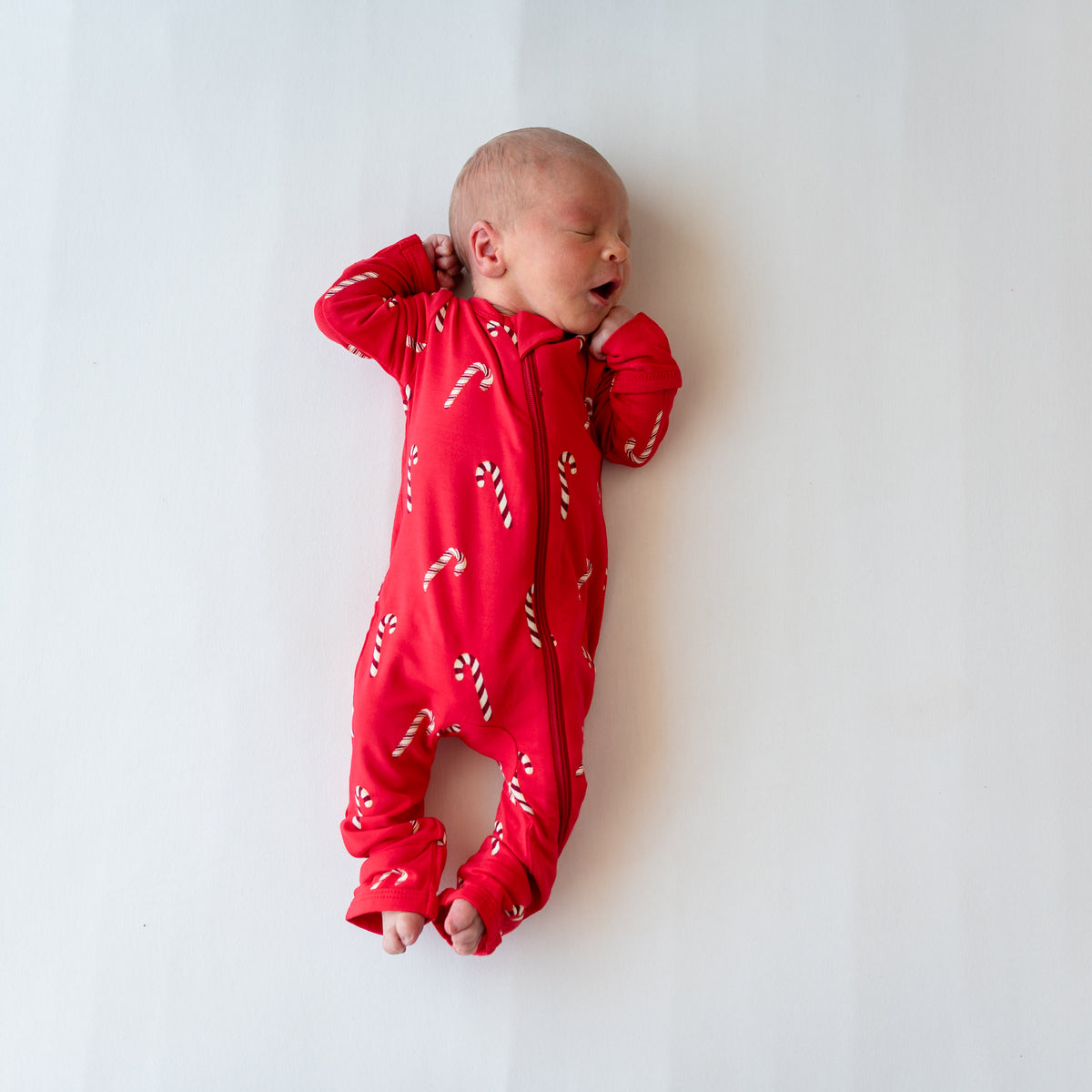 Sleeping newborn laying on a white blanket wearing the Zippered Romper in Candy Cane