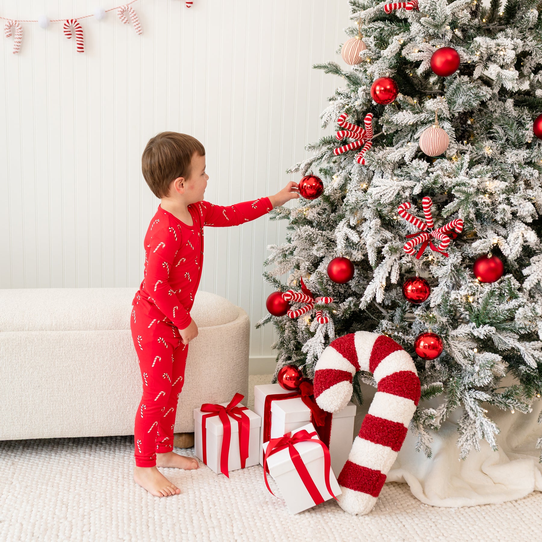 Young boy standing beside a decorated Christmas tree wearing the Long Sleeve Pajamas in Candy Cane touching one of the Christmas decorations on the tree