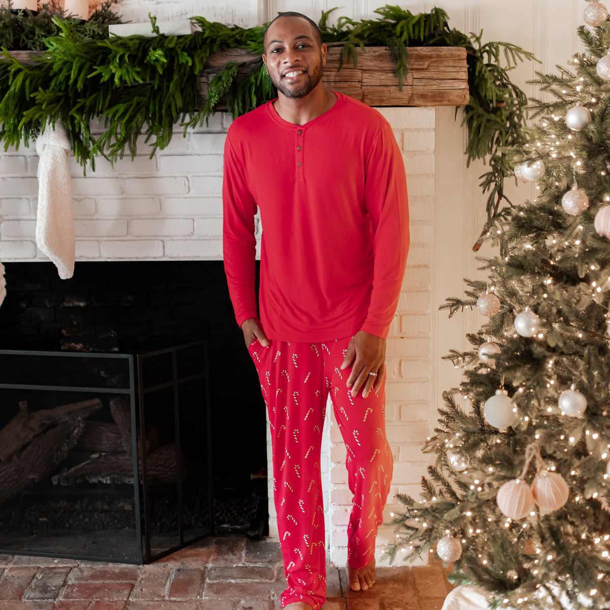 Male model standing in front of a white brick fireplace wearing the Men's Lounge Pants in Candy Cane and Men's Henley top in Cardinal