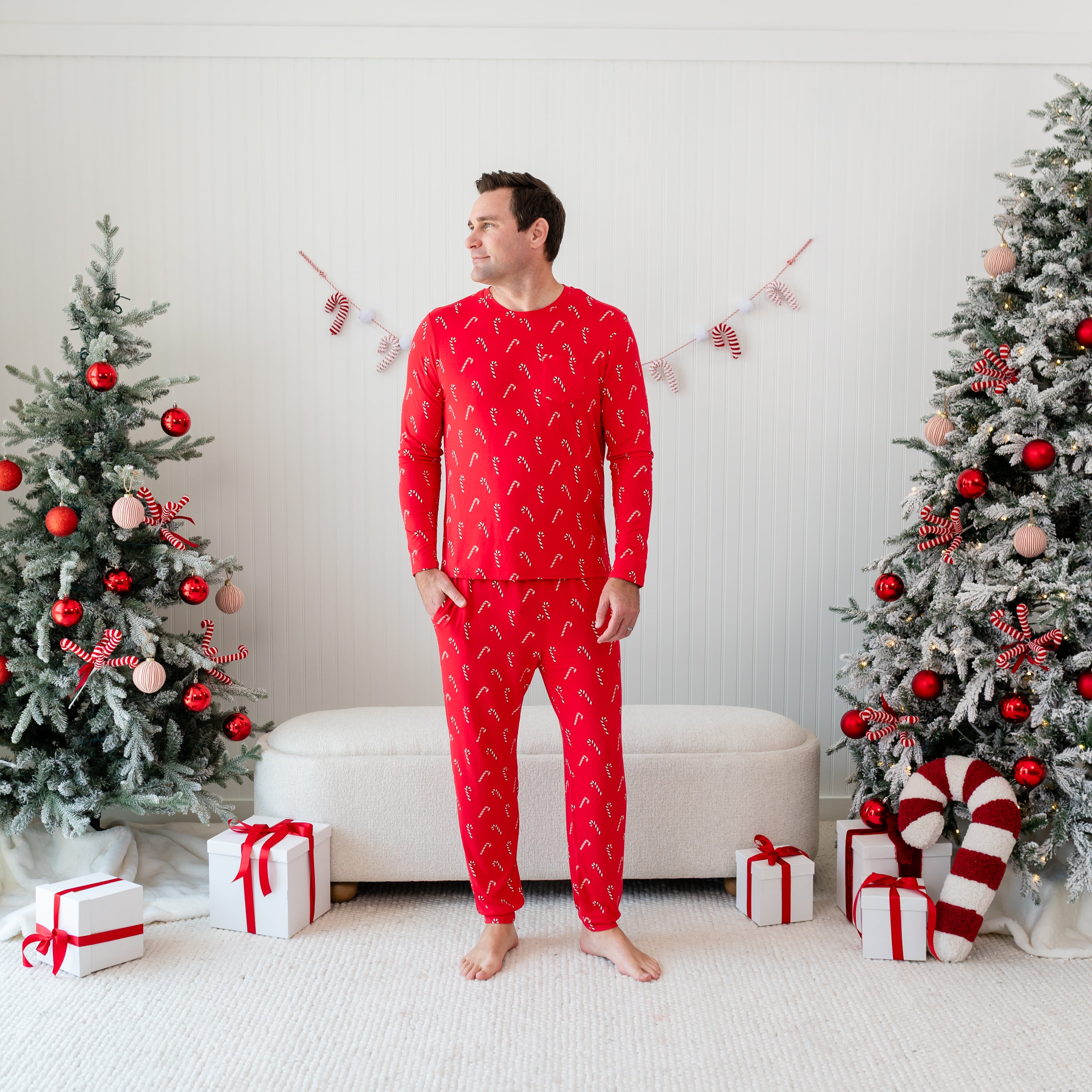 Male model standing with one hand in his pocket wearing the Men's Jogger Set in Candy Cane in front of a cream ottoman between two decorated Christmas trees