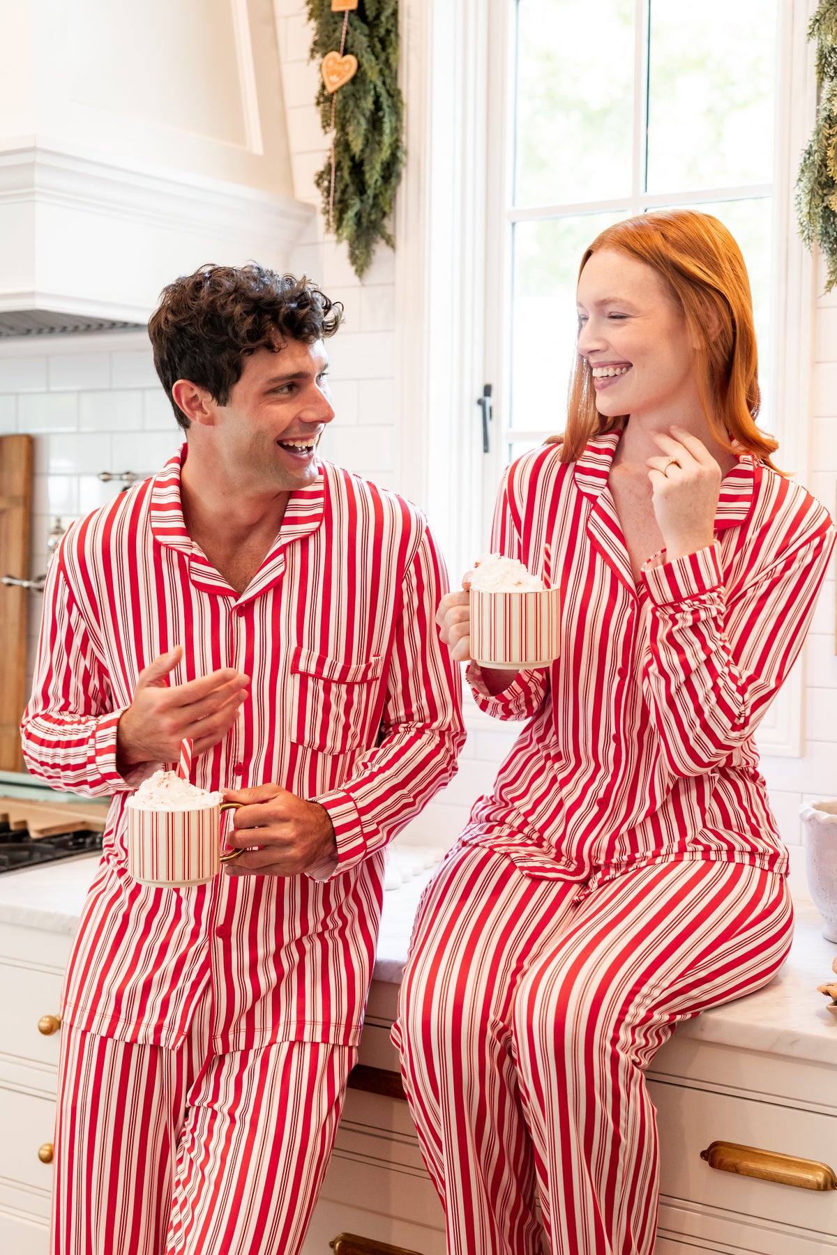 A man and a woman wearing matching red and white vertically striped pajamas sit on a kitchen counter, each holding a striped mug with whipped cream.