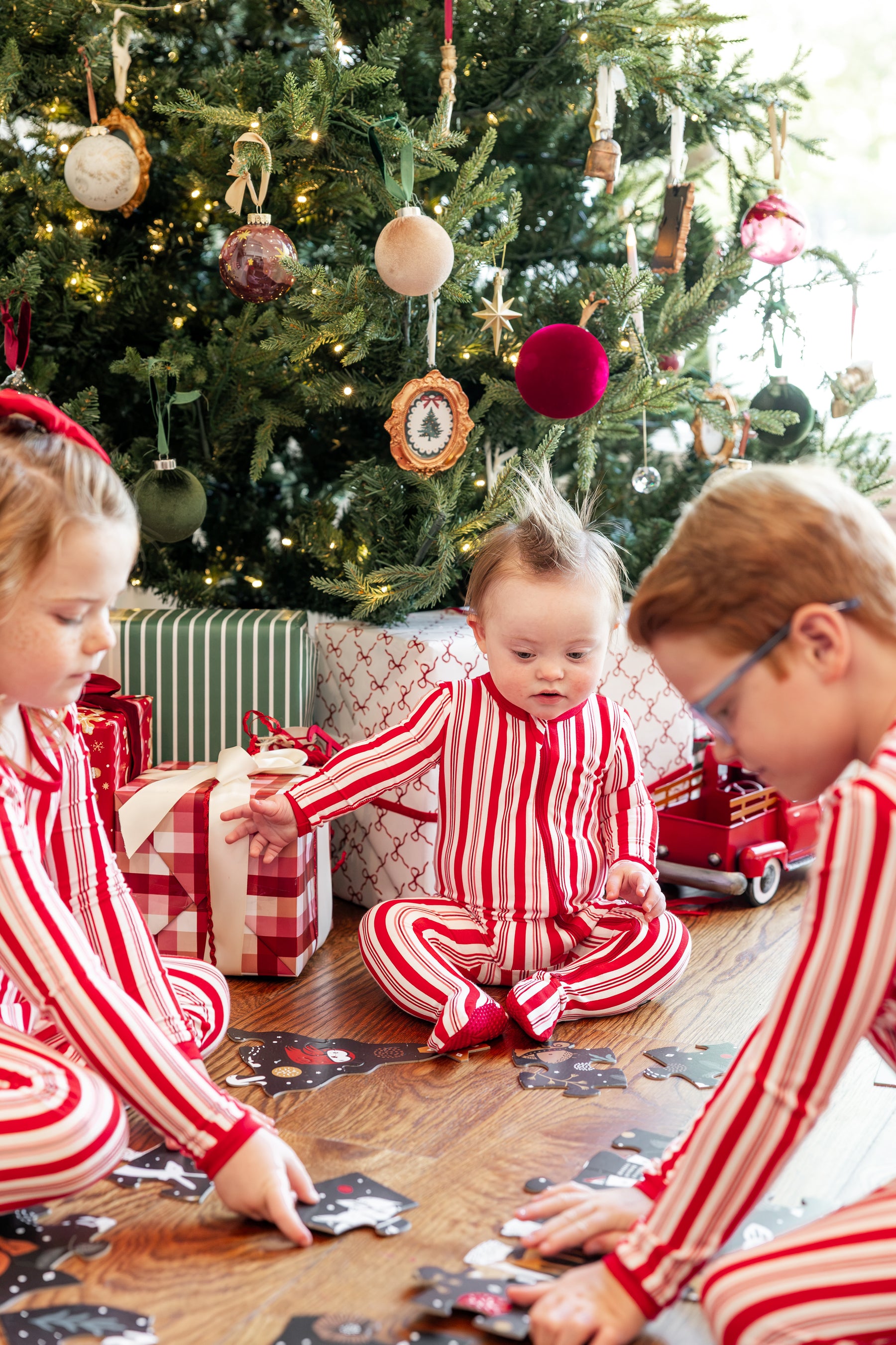 Three children wearing matching red and white striped Christmas pajamas sit on the floor in front of a decorated Christmas tree, with wrapped gifts and a red toy truck nearby.