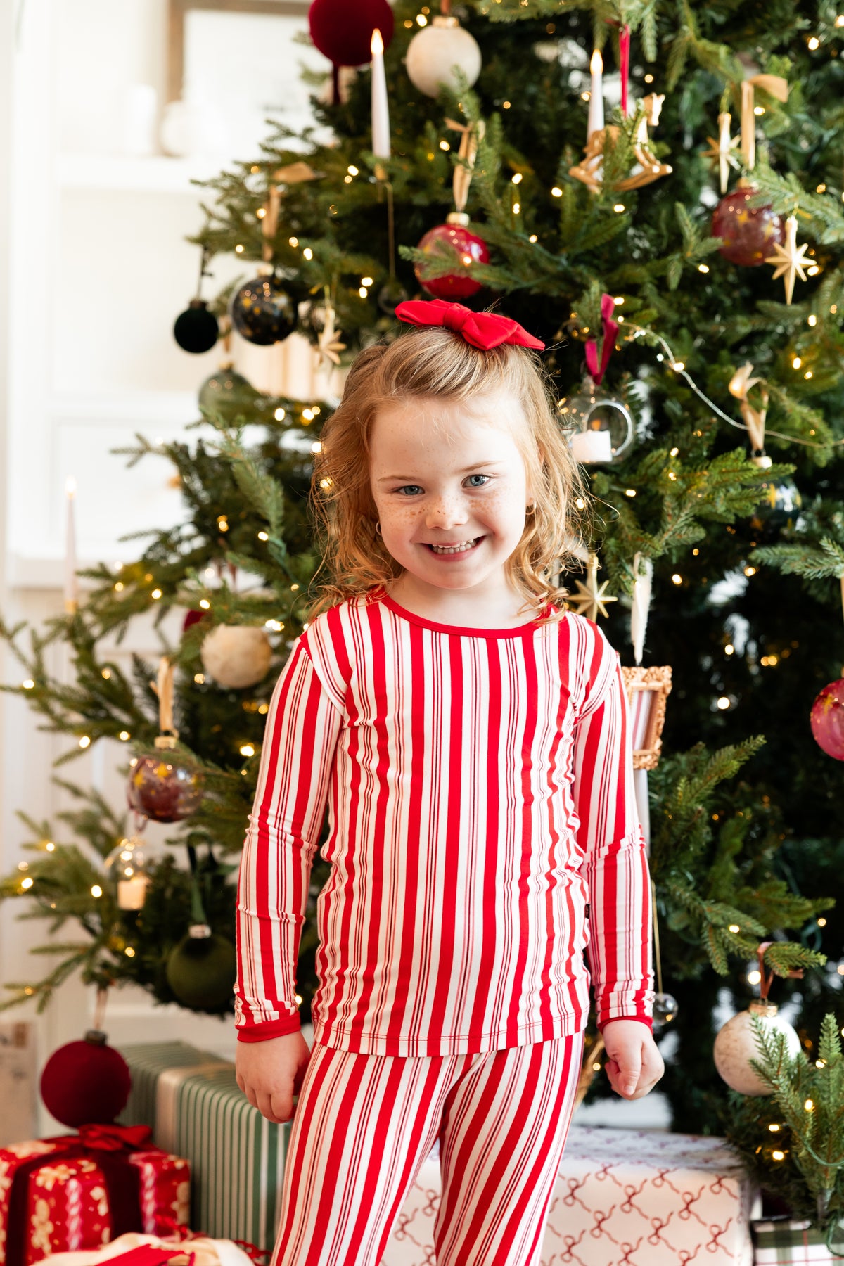 Girl wearing Long Sleeve Pajamas in Peppermint Stripes print and Red Bow with Christmas tree in the background