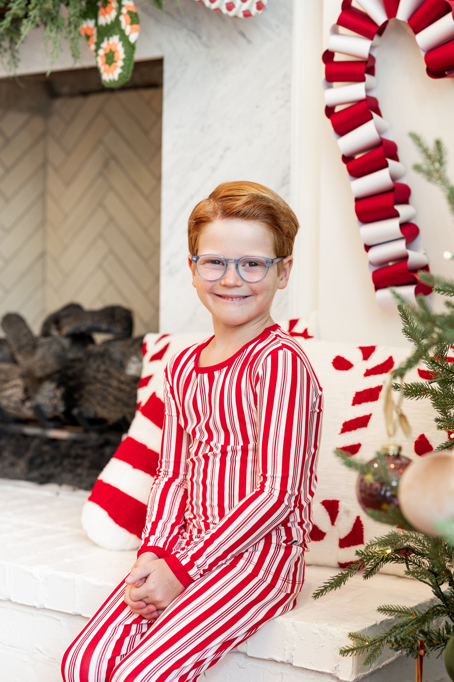 A young boy with red hair and glasses wearing red and white striped Christmas pajamas, sitting on a white fireplace mantel decorated with holiday garlands and a candy cane.