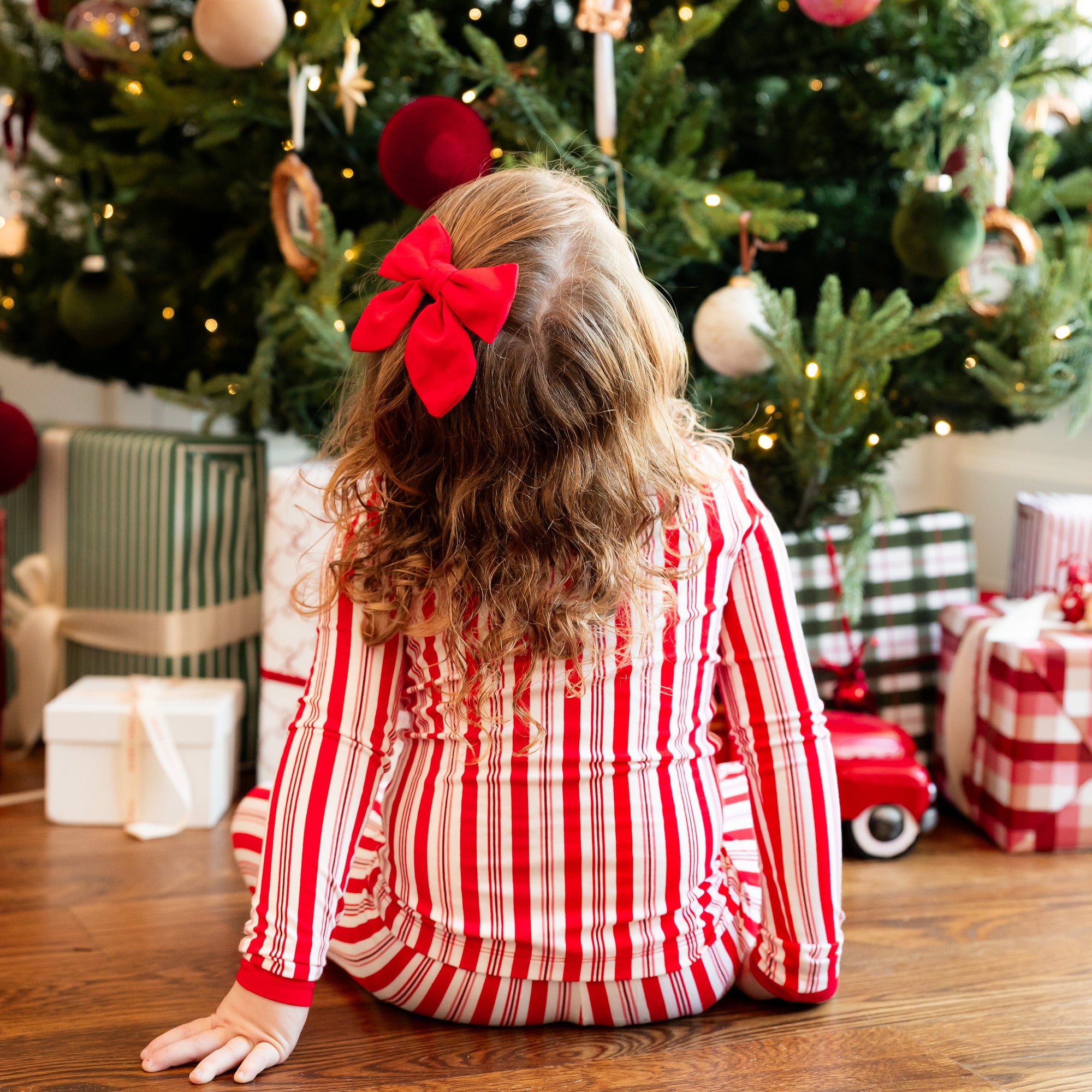 A young girl with a large red bow in her hair sits on the floor in front of a decorated Christmas tree, wearing a red and white striped pajama top. Gift boxes are wrapped around the tree, and a small red toy car is visible on the floor.