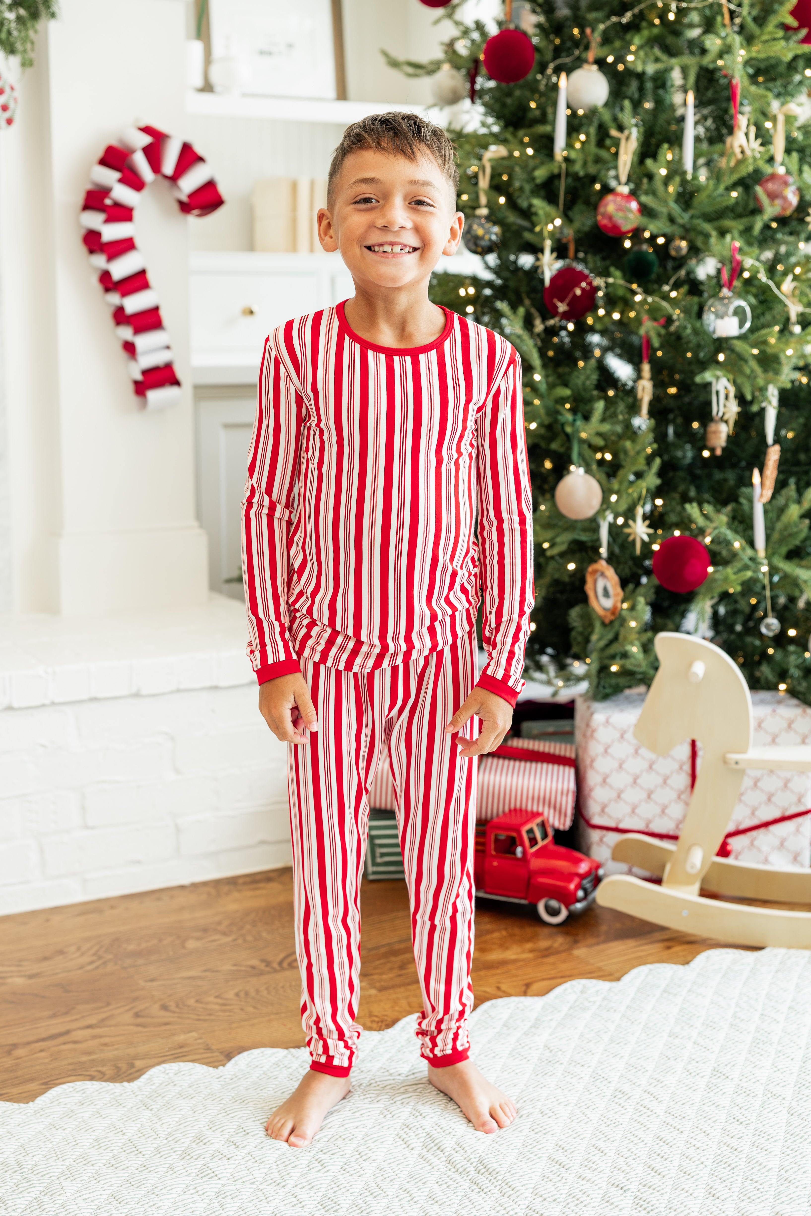 Boy wearing Long Sleeve Pajamas in Peppermint Stripes print with Christmas tree in the background