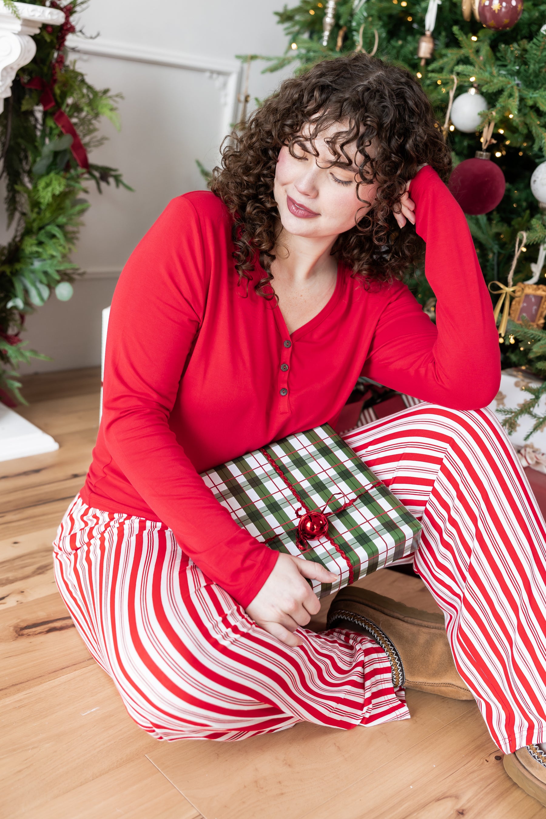 Modal sitting in front of Christmas tree with present wearing Women's Lounge Pants in red and white Peppermint Stripes print under a red T-shirt