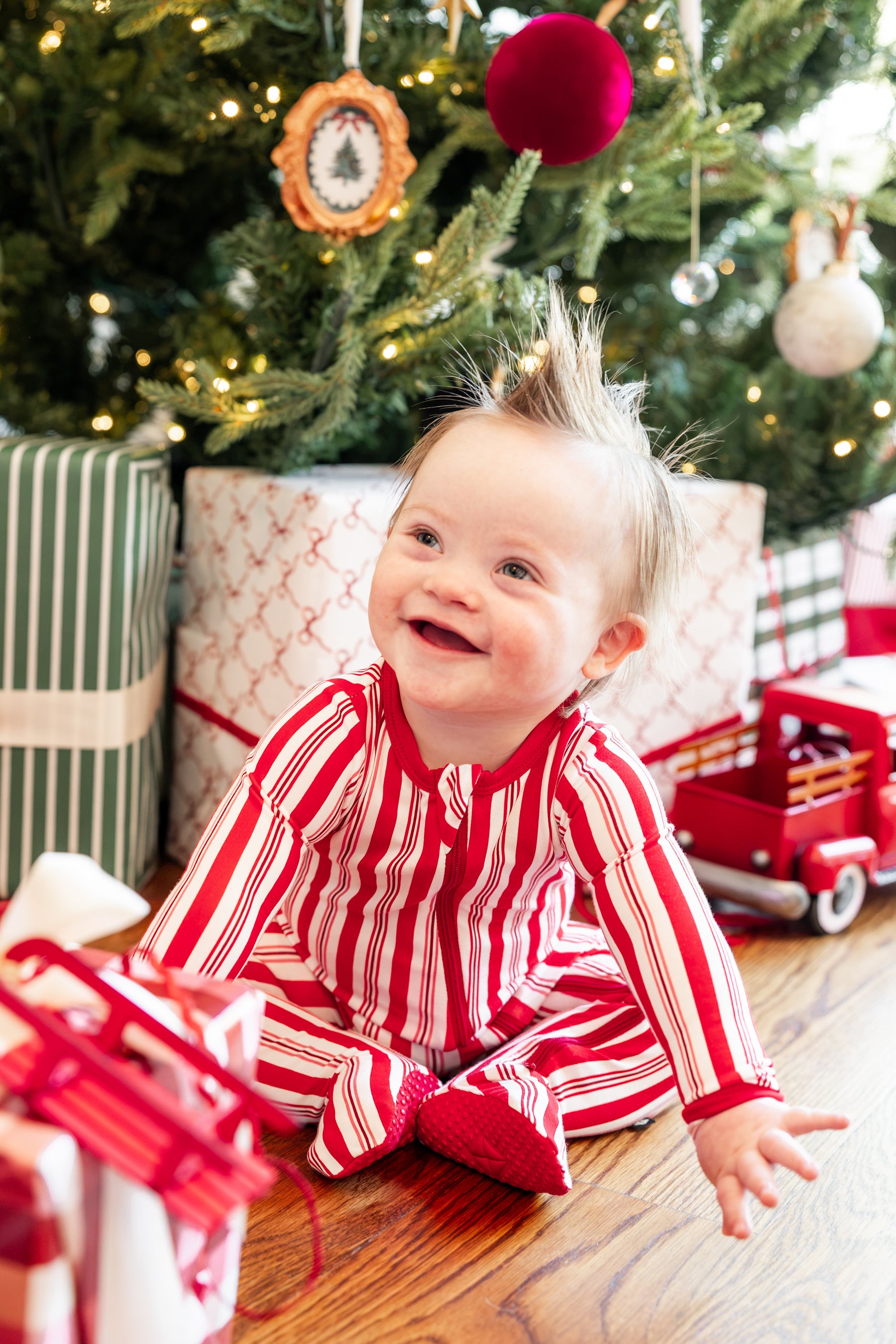 Baby wearing Zippered Footie in red and white Peppermint Stripes print  sitting with presents