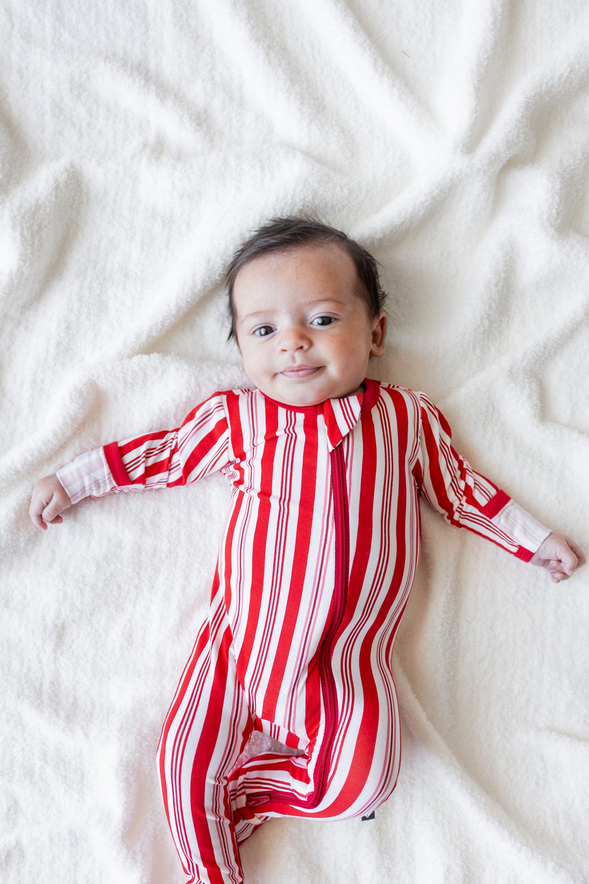 Baby wearing Zippered Footie in red and white Peppermint Stripes print lying on a white blanket