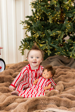 Baby wearing Zippered romper with red and white Peppermint Stripes print while sitting with Baby Doll Sleep Bag in  similar print on a blanket in front of a Christmas tree
