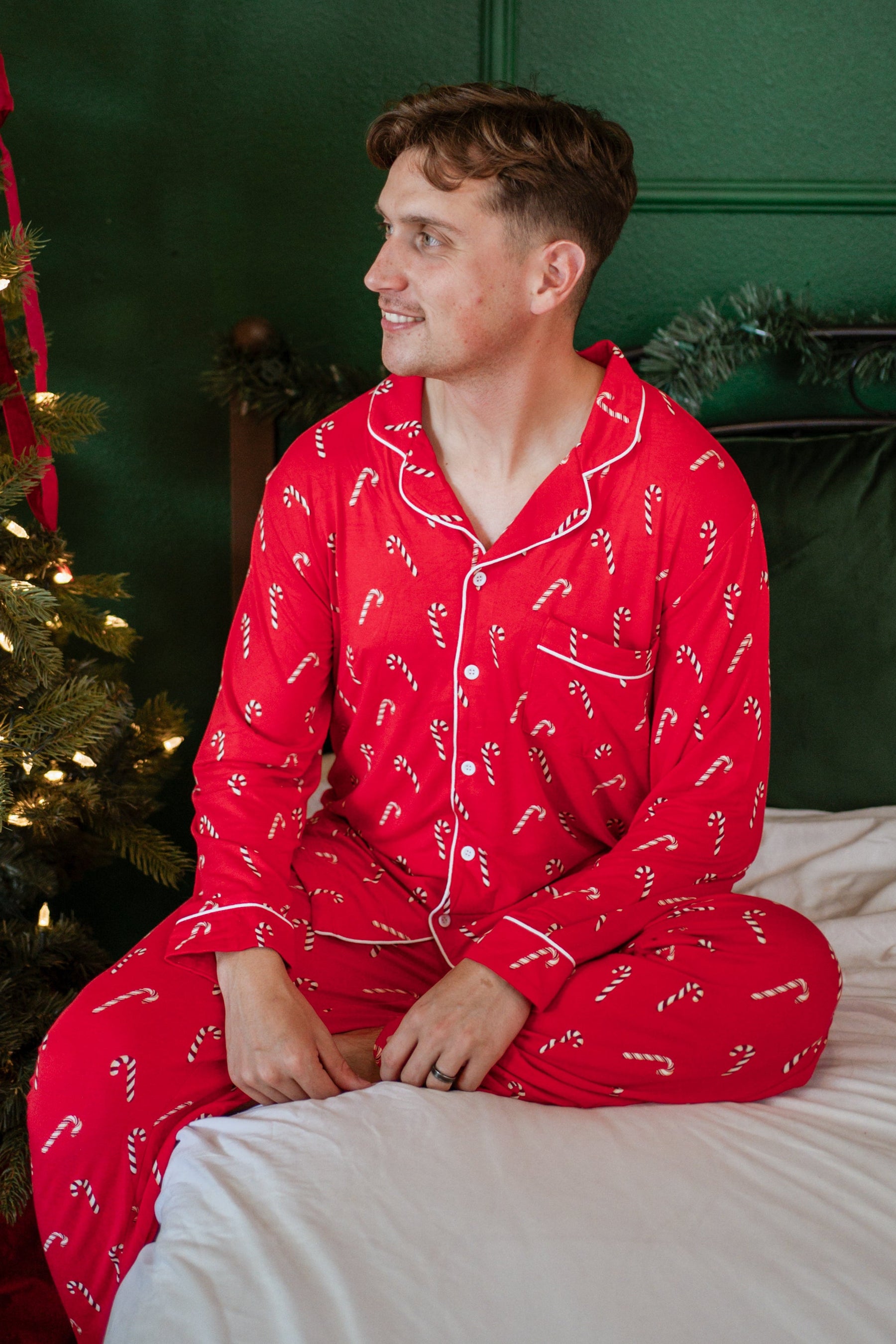 Male model sitting on a bed wearing the Men's Long Sleeve Pajama Set in Candy Cane with a decorated Christmas tree beside him