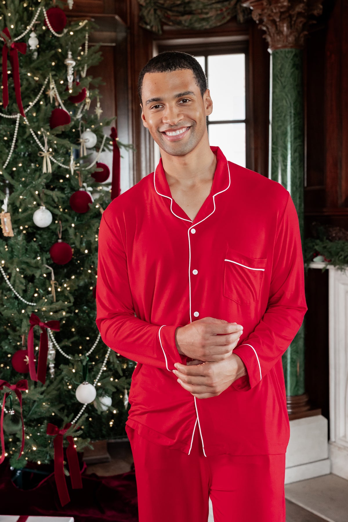 A man wearing a bright red long-sleeve pajama set with white piping stands in front of a decorated Christmas tree in an indoor holiday setting.