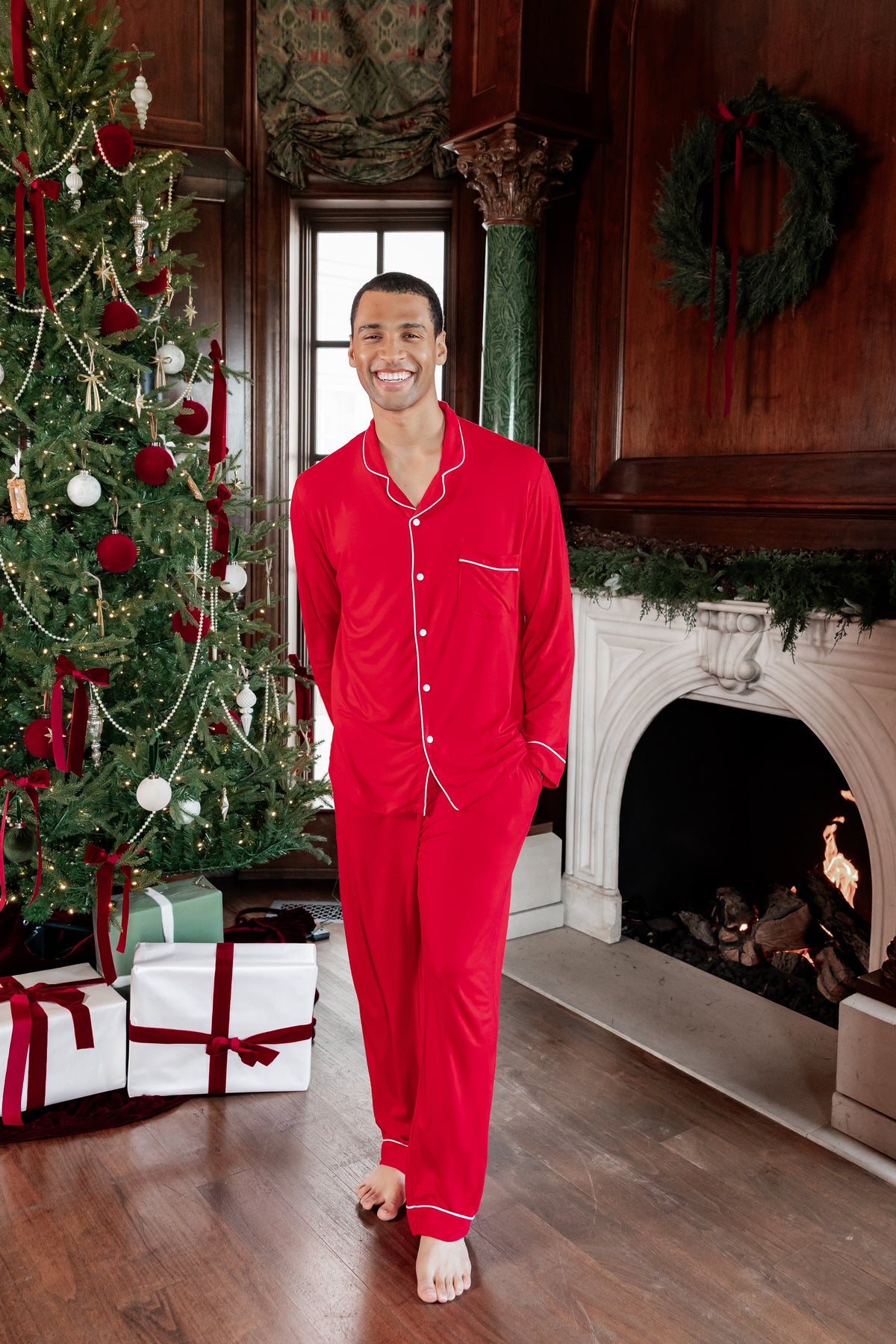 A man wearing a bright red two-piece pajama set with white piping stands barefoot in a cozy holiday living room with a decorated Christmas tree and fireplace.