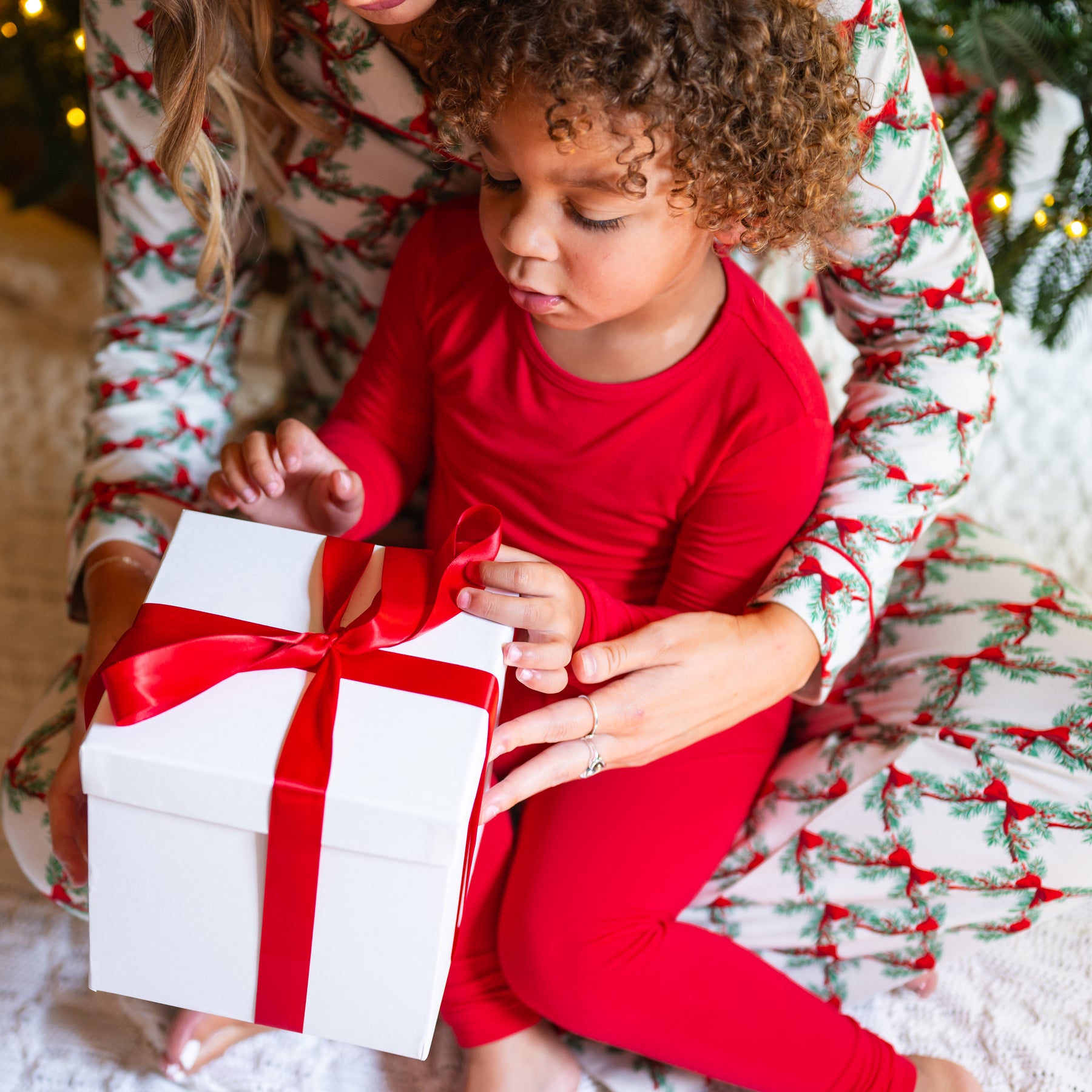 Young toddler sitting on mothers lap wearing the Long Sleeve Pajamas in Cardinal opening a gift