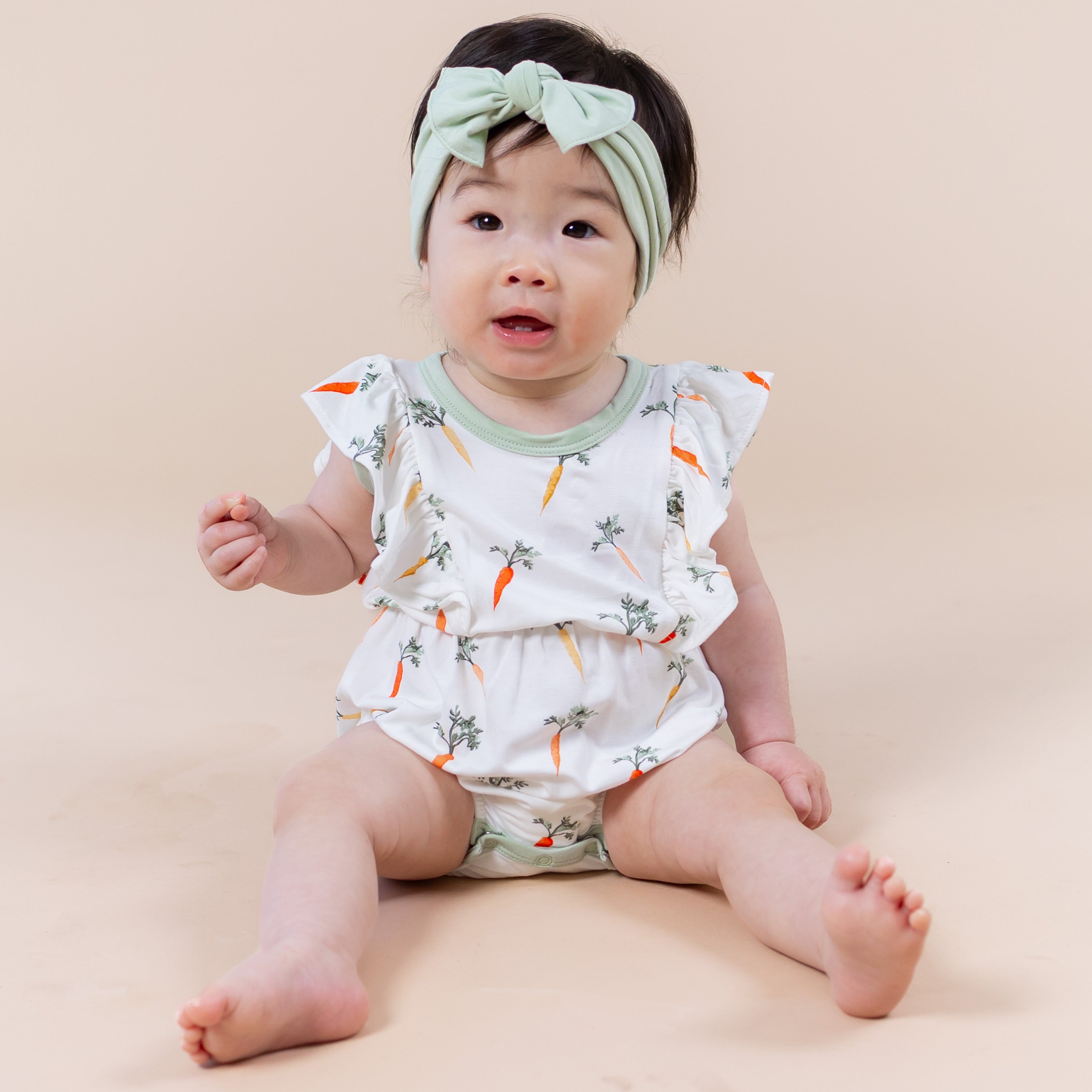 Young toddler sitting on the floor wearing the Bubble Romper in Carrot with green bow in her hair