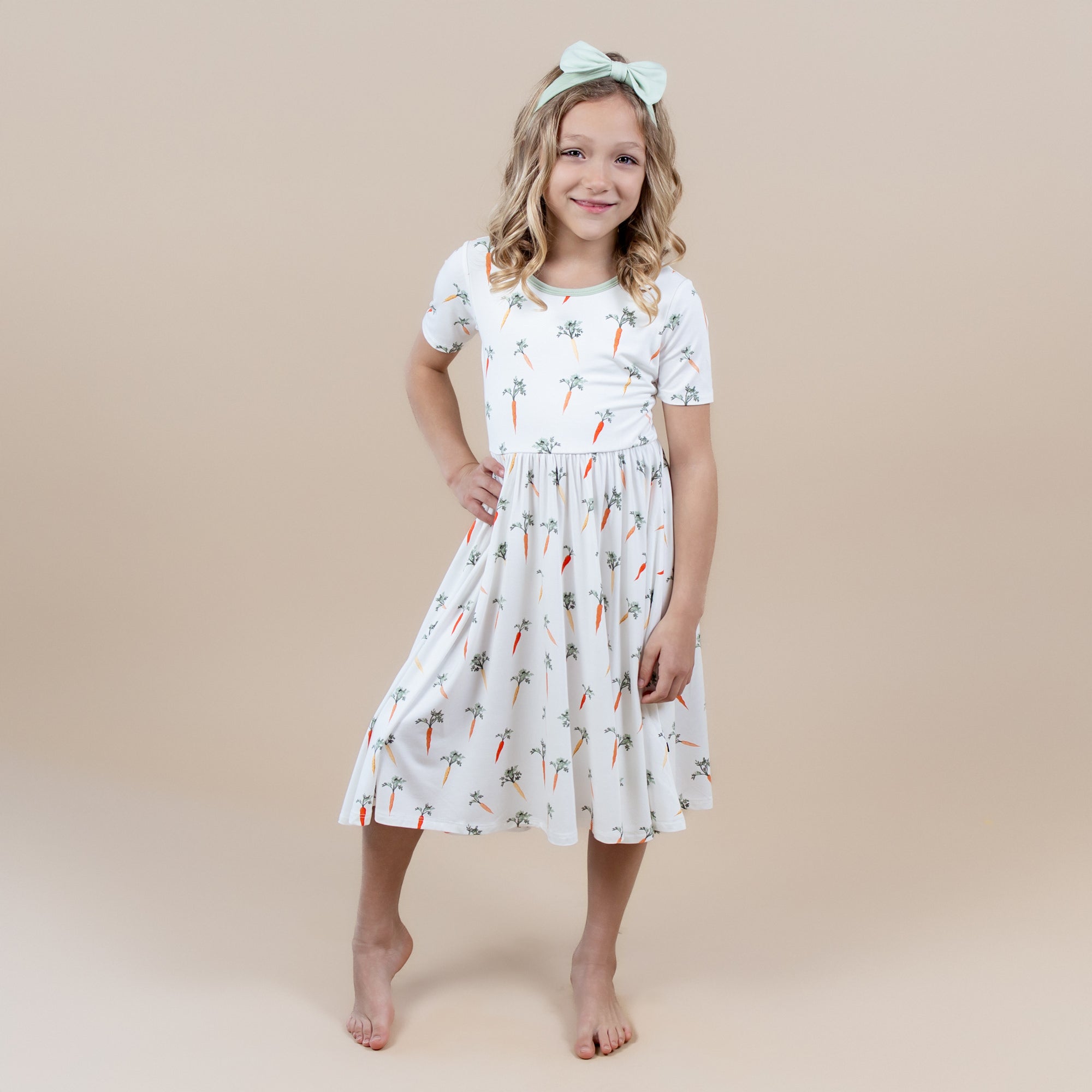 Young girl posing wearing the Twirl Dress in Carrot paired with a light green bow